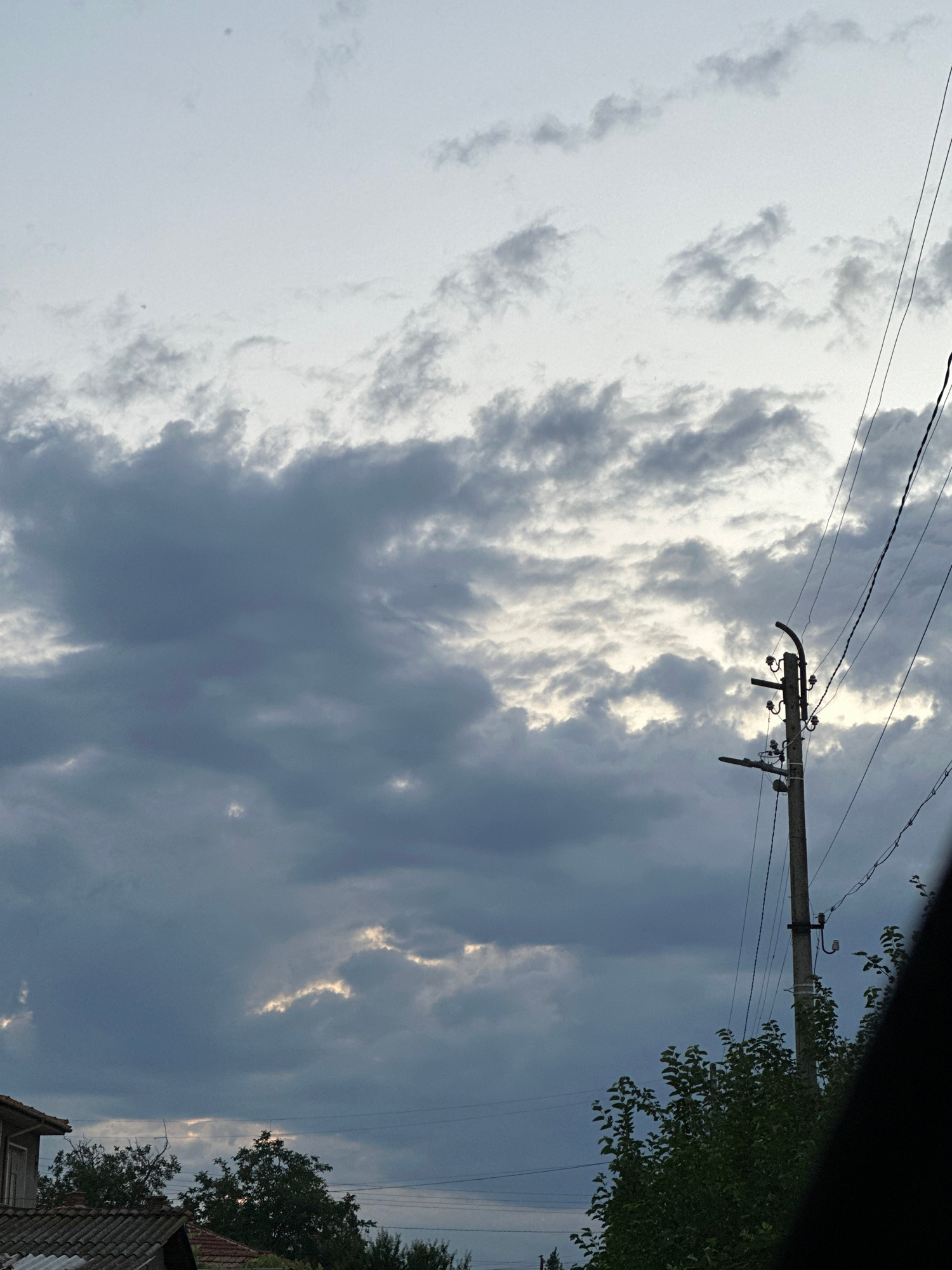 The image depicts an overcast sky with dramatic cloud formations, illuminated by breaks of sunlight. A telephone pole with electrical wires is prominent in the right-center of the frame. In the lower left, a portion of a tiled roof and some trees are visible, suggesting a residential area. The scene is likely located in or around Saedinenie, Bulgaria, given the architectural style of the roof and the presence of traditional utility poles. There are no people or visible animals in the image.