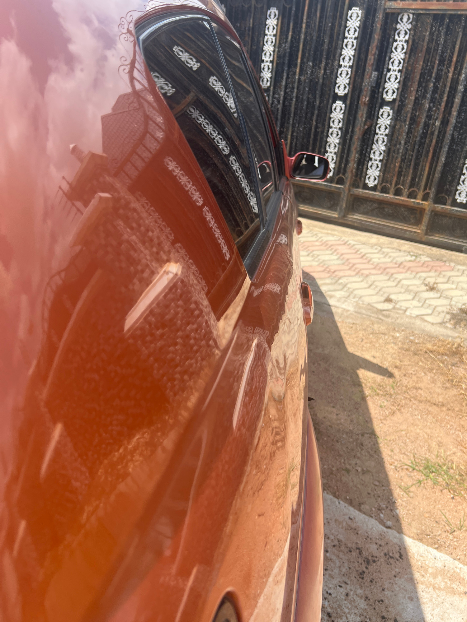 An orange car is parked parallel to a black wrought-iron gate. The car's surface reflects a building with a tiled roof and cloudy sky. The gate has decorative white patterns. The ground is a mix of concrete pavers and sandy dirt. The scene is outdoors, likely in an urban or suburban area in Howayi, Nigeria.