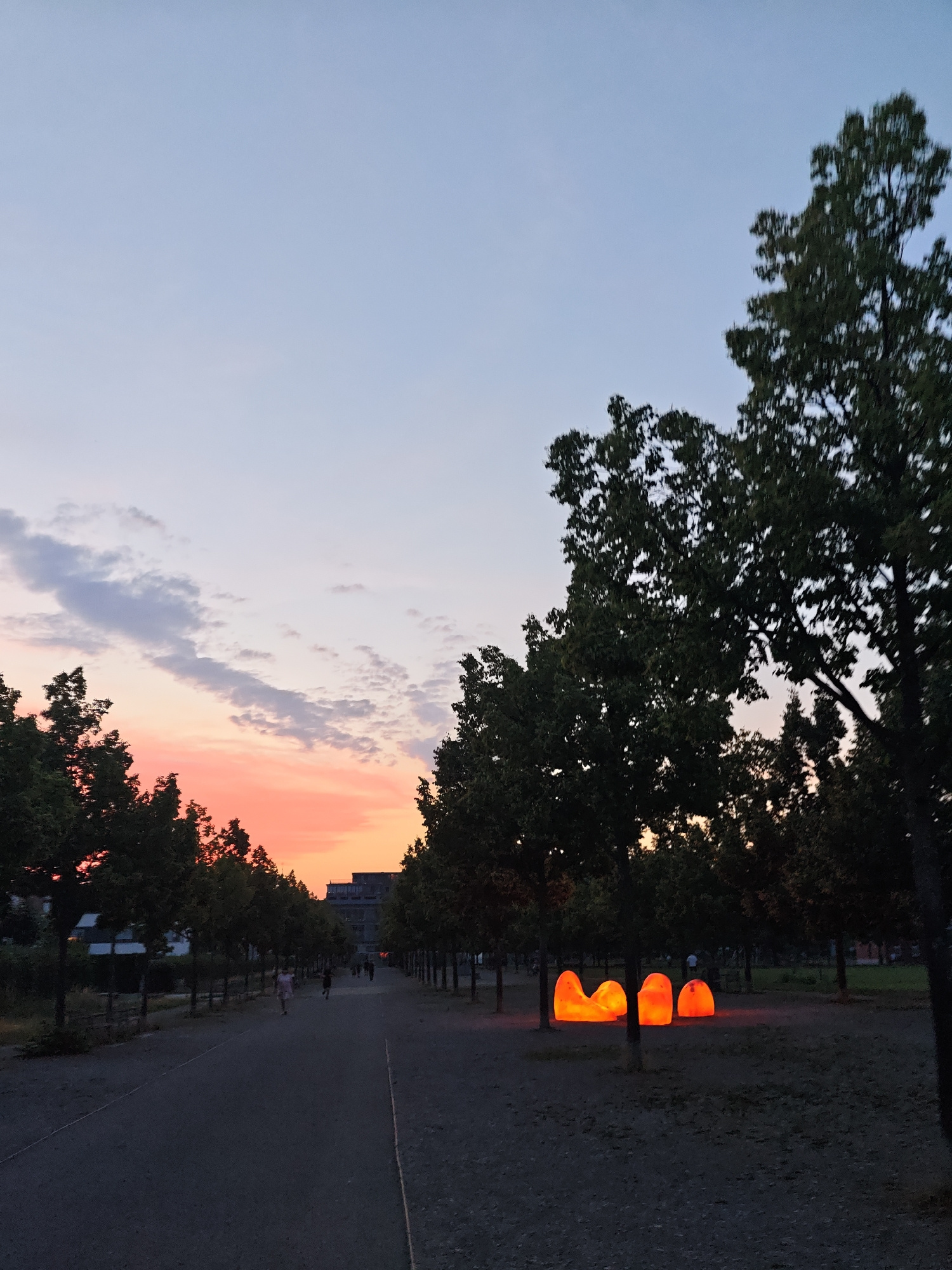 The image captures a park setting in Fürth, Germany, during twilight. A pathway, lined with trees on both sides, stretches into the distance. Several illuminated, amorphous orange sculptures are visible on the right side of the path, nestled amongst the trees. The sky displays a gradient of soft blues and pinks from the setting sun, with wispy clouds. In the background, a building complex is silhouetted against the vibrant sky. A few individuals are walking or standing along the pathway in the mid-ground, while others are further in the distance. The ground is covered in gravel or fine dirt.
