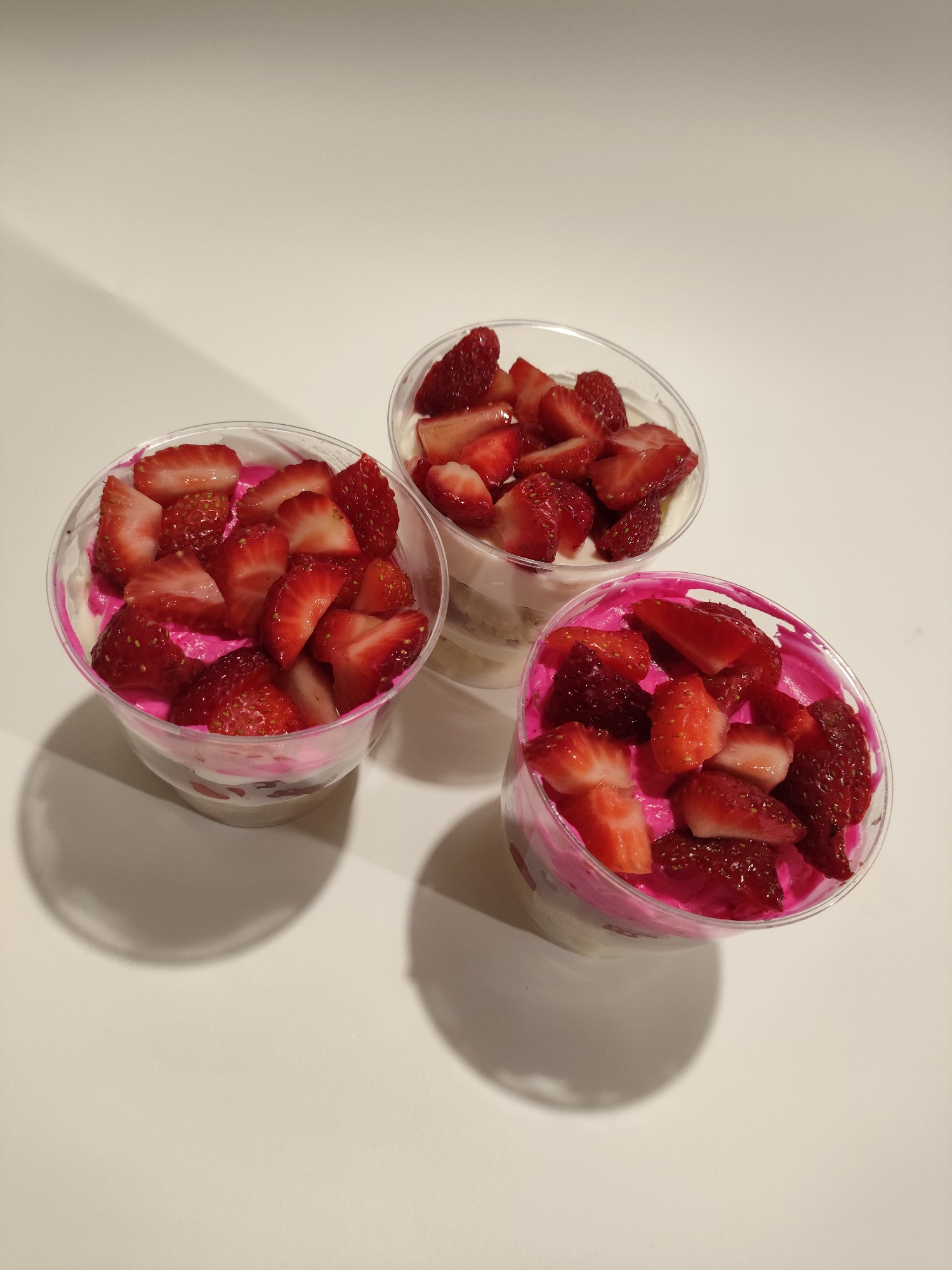 Three clear plastic cups filled with a dessert are arranged on a white surface. Each cup contains layers of white cream and sliced strawberries. One cup has a vibrant pink layer visible beneath the strawberries, while the other two have white cream only. The lighting creates shadows on the surface. The image does not contain any elements that would allow for the identification of a location in Myadzyel, Belarus.