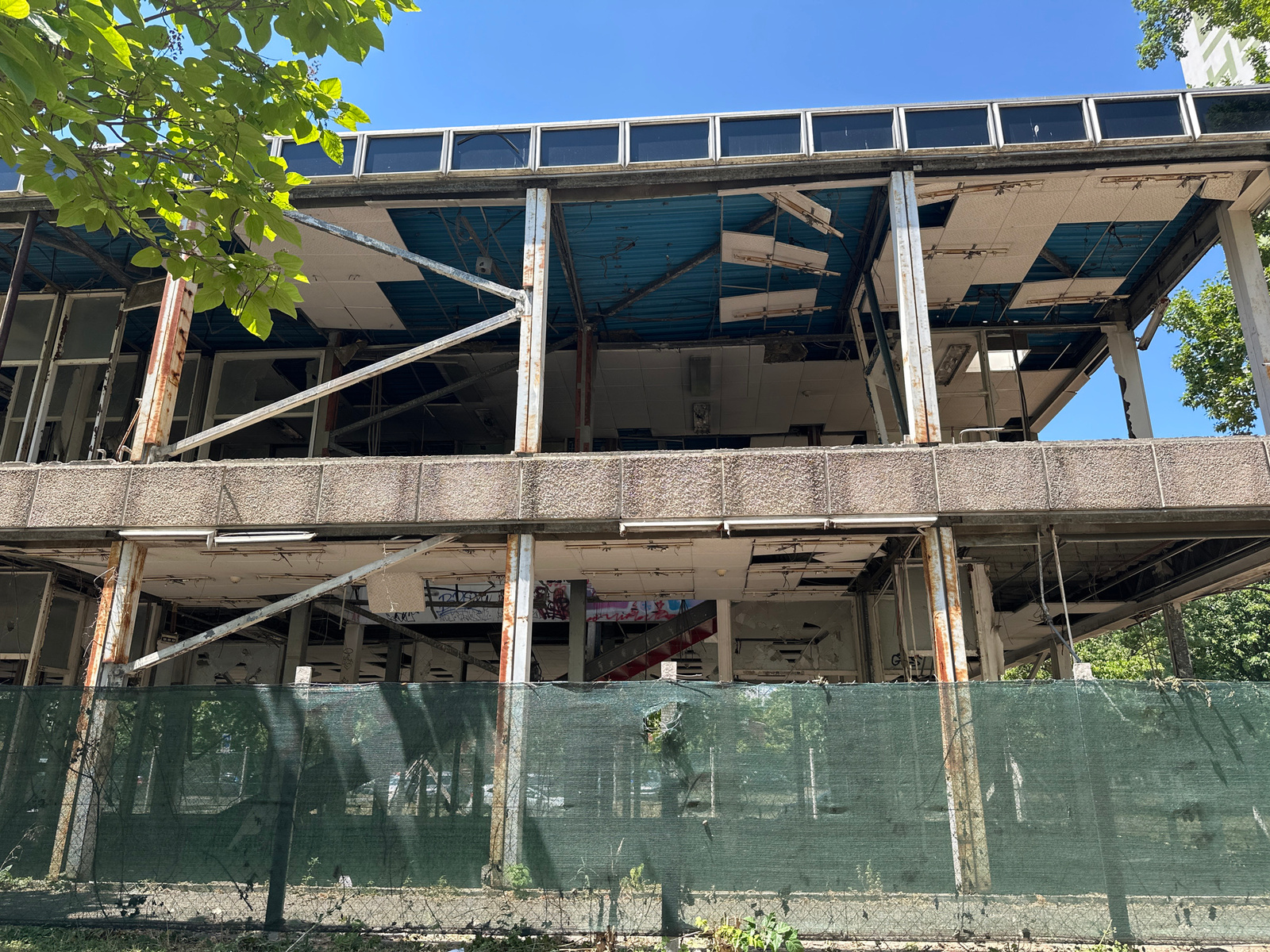 The image shows a dilapidated multi-story building, likely undergoing demolition or in a state of severe disrepair. The facade is concrete with a textured finish, and large sections of the building's interior are exposed, revealing a skeletal structure of metal beams and concrete floors. The windows are mostly broken or missing, and the ceilings are damaged, with hanging wires and loose ceiling tiles visible. Some graffiti can be seen on the interior walls. A green mesh fence runs along the front of the property, obscuring the ground level and indicating a restricted or hazardous area. Overgrown vegetation and weeds are present in the foreground and around the fence. The sky is clear and blue, suggesting a sunny day. The overall impression is one of abandonment and decay. The image's context is Budapest, Hungary, but specific landmarks are not identifiable. No people are visible in the image, but the scene suggests a recent or ongoing deconstruction event.