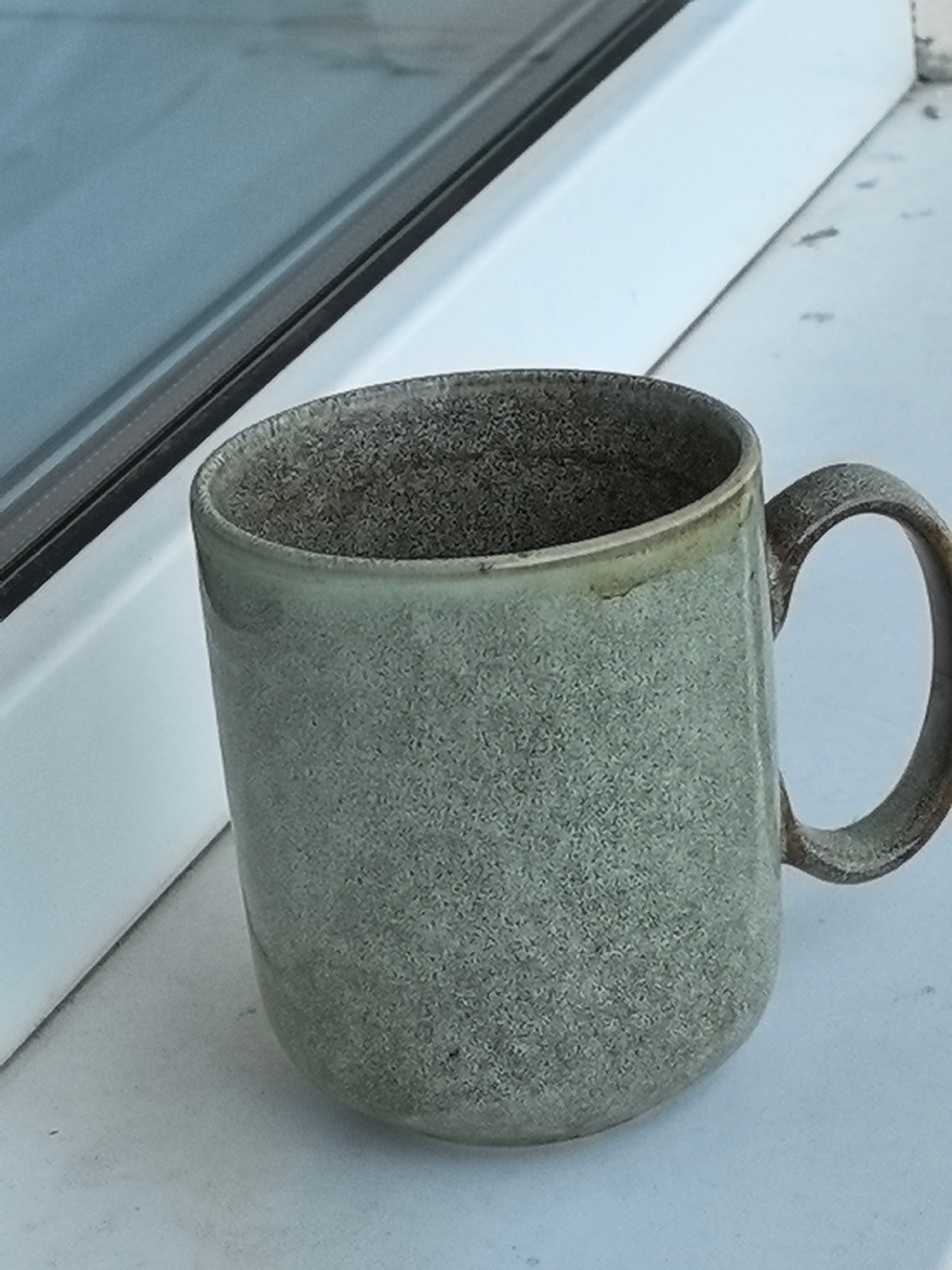 A close-up, eye-level, medium shot captures a speckled, light green ceramic mug with a textured handle, positioned on a white ledge. The mug is oriented with its handle to the right. Behind the mug, a blurred, blue-tinted window frame or surface is visible, with a thin black line running horizontally. The white ledge appears to be dusty, with small dark specks scattered across its surface. The lighting suggests an indoor environment with natural light coming from the left.