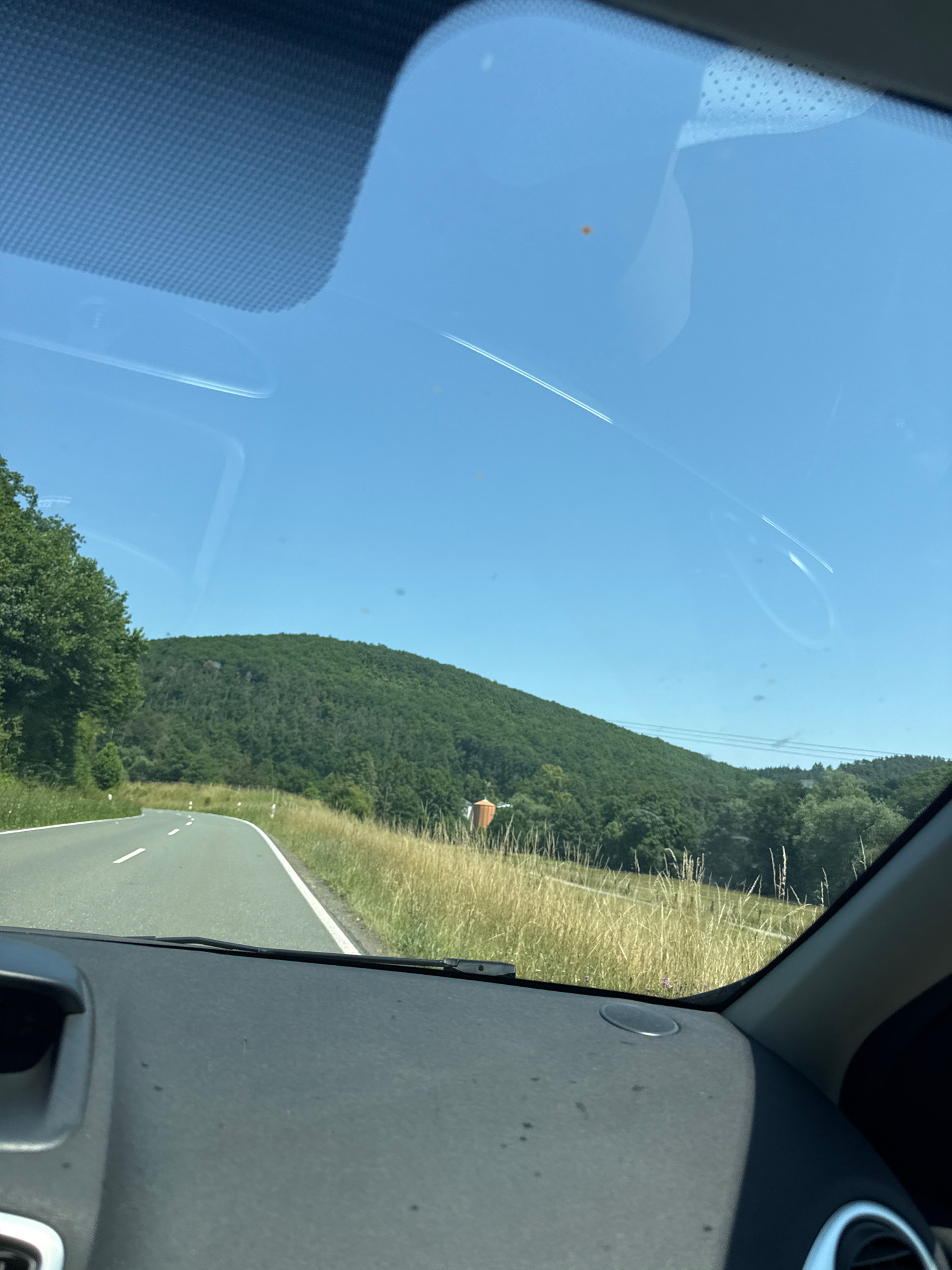 This image depicts a view from inside a vehicle, looking forward through the windshield on a clear, sunny day. The road ahead curves to the left, bordered by tall, dry grass on the right and trees on the left. In the background, a lush green, tree-covered hill dominates the landscape. Faint contrails are visible in the bright blue sky. On the right side of the frame, part of the vehicle's dashboard is visible, along with a portion of the car's interior trim. A small, round, orange structure, possibly a water tower or silo, is situated on the slope of the hill in the mid-ground. The scene is consistent with a rural setting in or near Nauborn, Germany, characterized by rolling hills and agricultural or forested areas.