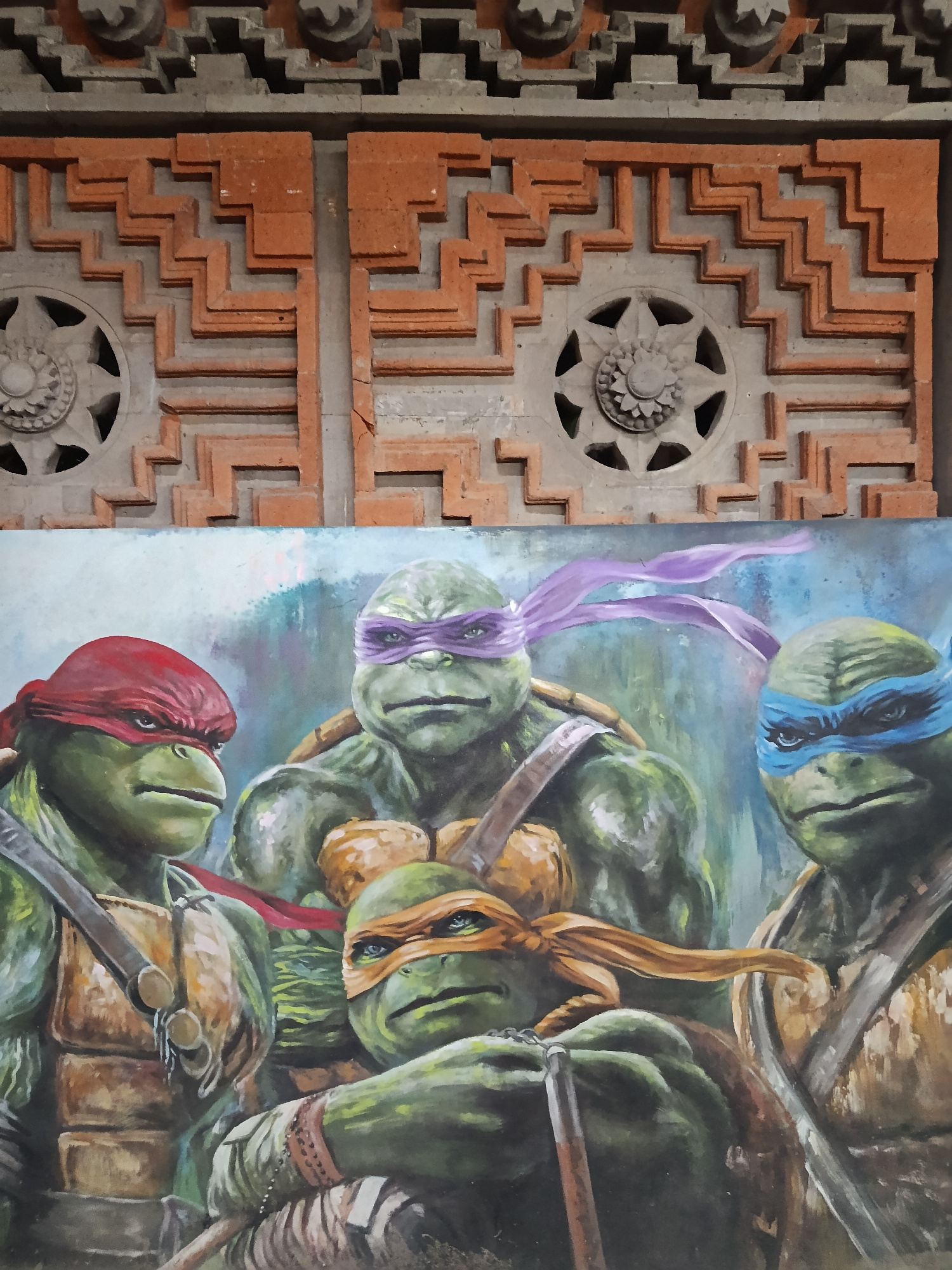 The image displays a detailed, vibrantly colored oil painting of the Teenage Mutant Ninja Turtles, set against a textured background of ornamental brickwork. The four turtles are depicted in close proximity, with their iconic colored bandanas clearly visible: Raphael in red, Donatello in purple, Leonardo in blue, and Michelangelo in orange. They are shown with serious expressions, their muscular forms rendered with visible brushstrokes. The brickwork above the painting features intricate, geometric patterns and circular decorative elements, suggesting an architectural or decorative facade. The overall scene combines a piece of art with its surrounding environment, implying it is a mural or a painting displayed on a wall.