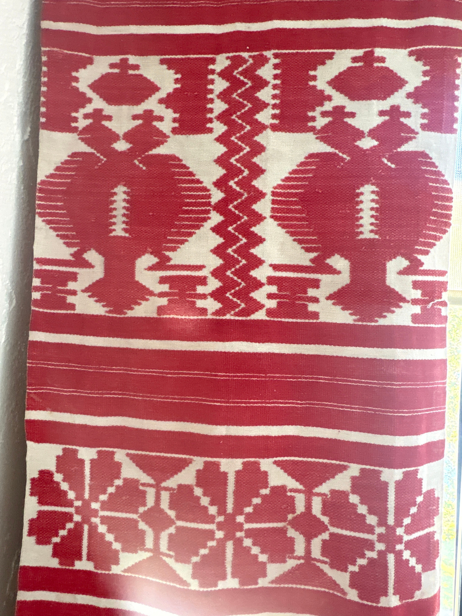 A red and white woven textile is displayed vertically. The textile features geometric patterns. At the top, there are two stylized bird or eagle-like figures in red on a white background, separated by a zig-zag pattern. Below these figures are horizontal red and white stripes. Further down, the textile shows repeating floral or starburst motifs in red on a white background, also bordered by red and white stripes. The textile appears to be hanging against a white textured wall. The environment is indoors, with natural light entering from the right, casting shadows on the textile. There are no people or animals present. The textile's origin or exact location in Delray Beach, United States of America, is not ascertainable from the image.