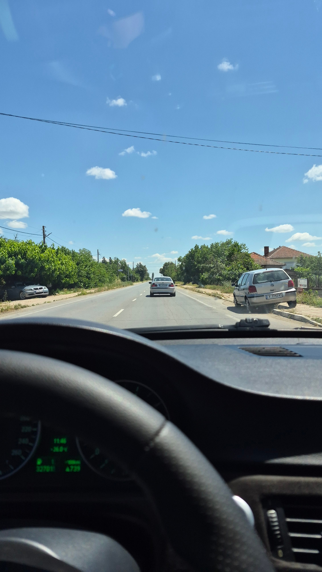 The image is taken from the driver's perspective inside a car, looking forward. The steering wheel is visible in the foreground, slightly blurred. Beyond the steering wheel, the road stretches ahead, with two lanes of traffic in each direction, separated by a white line. There are several cars on the road. In the distance, a silver sedan is traveling in the same direction as the viewer. To the right of the road, a silver hatchback is parked on the shoulder, partially on the curb. On the left side of the road, a dark grey coupe is parked under some trees. The surroundings are green and lush, with trees lining both sides of the road. The sky is a clear blue with scattered white clouds. There are power lines running overhead. The time is displayed on the car's dashboard as 11:46, with a temperature reading of -26.0°C and a mileage of 32701.