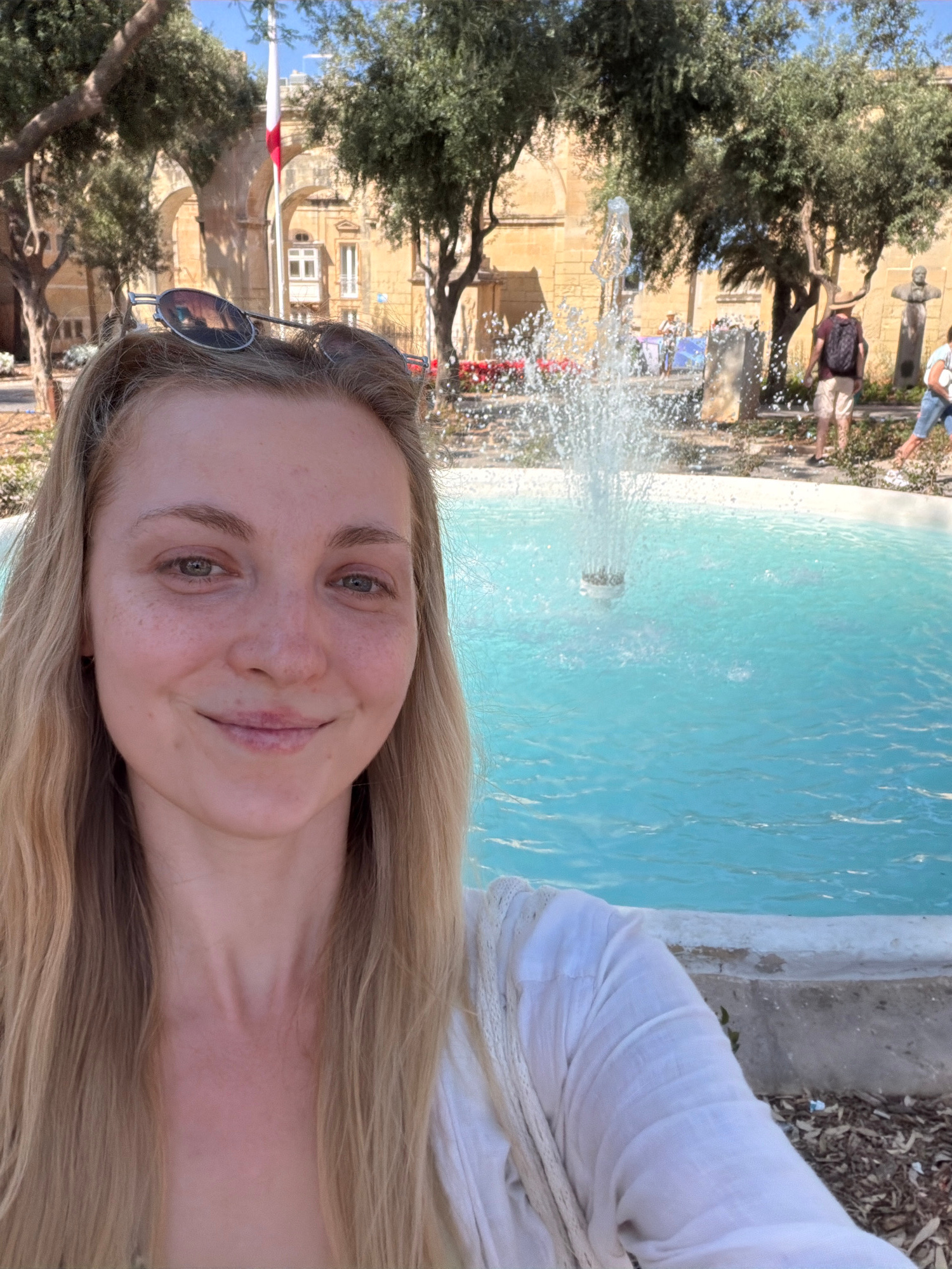 This media file is a portrait-oriented photograph featuring a young woman taking a selfie in front of a fountain. The woman has long blonde hair and is smiling, with sunglasses resting on her head. She is wearing a white top, and her expression suggests a happy and relaxed mood.
The background depicts an outdoor scene with a bright blue fountain in the foreground, actively spraying water. Behind the fountain, there are trees with lush green foliage and a historic building constructed of light-colored stone. The architecture of the building, with its arched entrances and windows, hints at a Mediterranean or European location. The presence of a flagpole with a red and white flag, and the characteristic stone buildings, strongly suggest the location is Valletta, Malta. The sunlight and the clear blue water indicate a bright, sunny day, likely during daytime. In the distance, a few other people can be seen, one of whom is wearing a backpack. The overall setting appears to be a public square or park, likely a tourist attraction given the historical architecture and fountain. There is no visible text in the image.