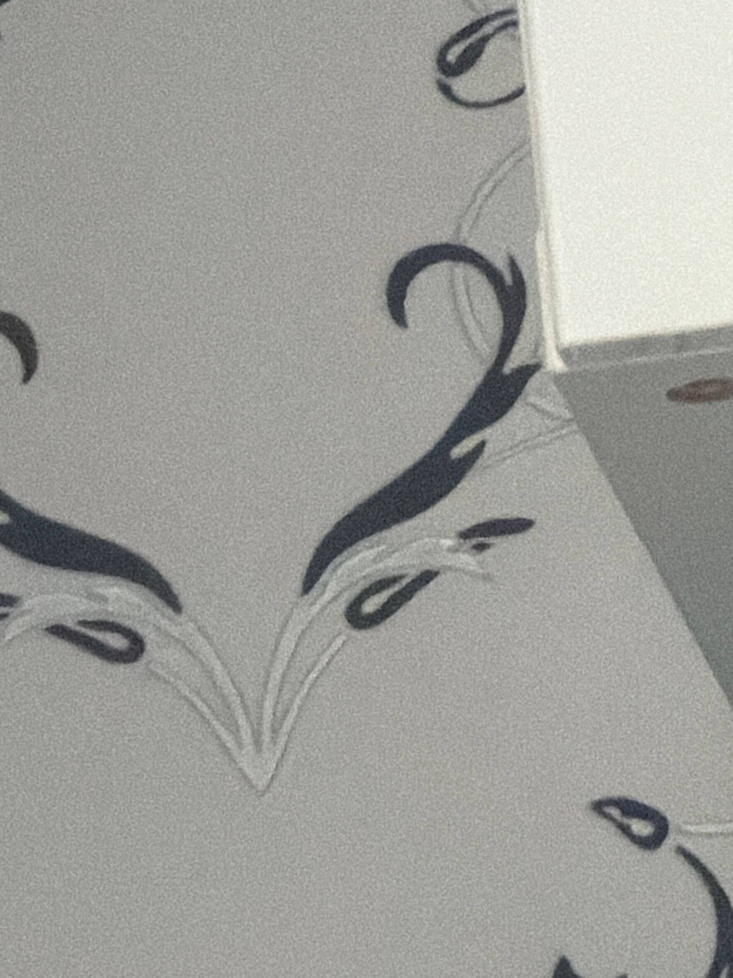 The image shows a close-up view of a wall with a patterned wallpaper. The pattern consists of dark blue, abstract floral or swirling shapes against a light gray, textured background. In the upper right corner, a portion of a white object, possibly a box or a wall extension, is visible. The lighting is soft, and the image has a grainy texture. There are no people or animals in the image. The scene appears to be indoors, in a domestic or commercial setting. The location is unknown.