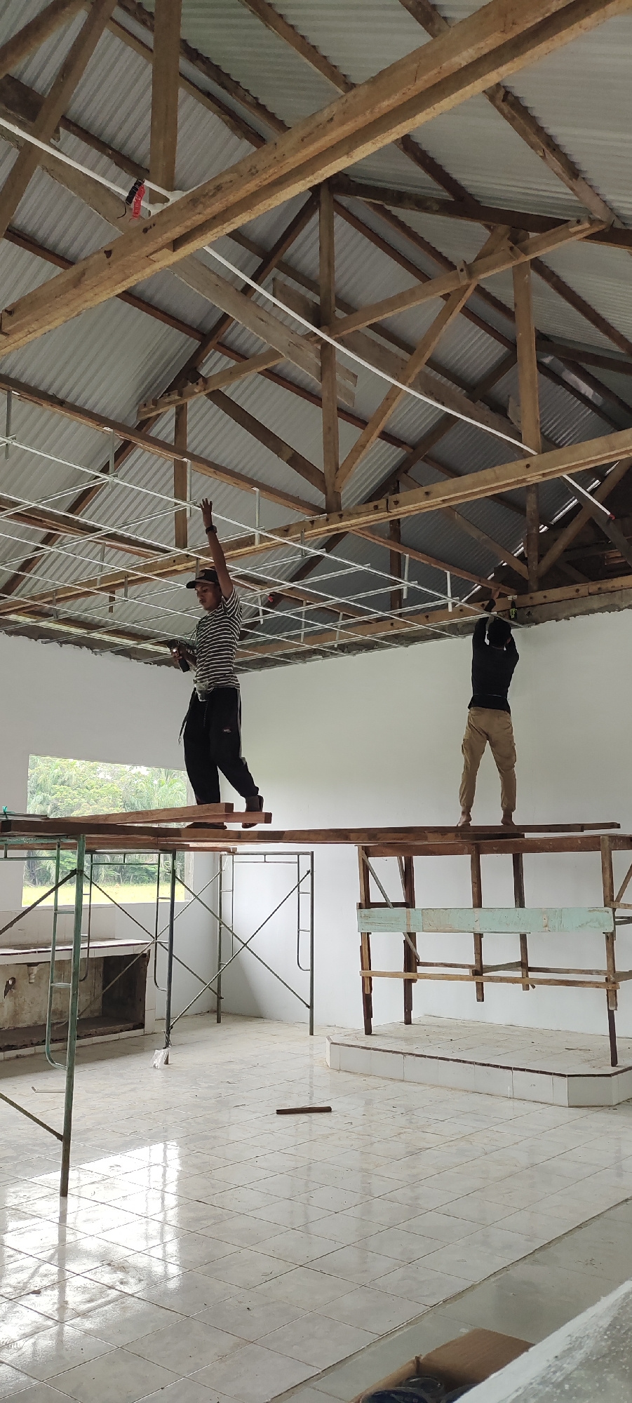 Two individuals are actively engaged in construction work inside a building with an exposed, wooden-raftered ceiling. The ceiling is partially covered with corrugated metal sheeting. One person, wearing a striped shirt, dark pants, and a baseball cap, stands on a wooden plank elevated by scaffolding. This individual is reaching upwards with their right hand, seemingly in the process of installing or adjusting something on the ceiling's metal grid system. The other person, dressed in a dark long-sleeved shirt, khaki pants, and a hat, stands on a similar wooden platform and is also facing the ceiling, with their back partially to the camera, appearing to be working on the ceiling grid as well. The environment is an unfinished interior space with white walls, a tiled floor, and a large window on the left wall offering a view of greenery. Various construction materials, including wooden planks and scaffolding, are visible in the scene.