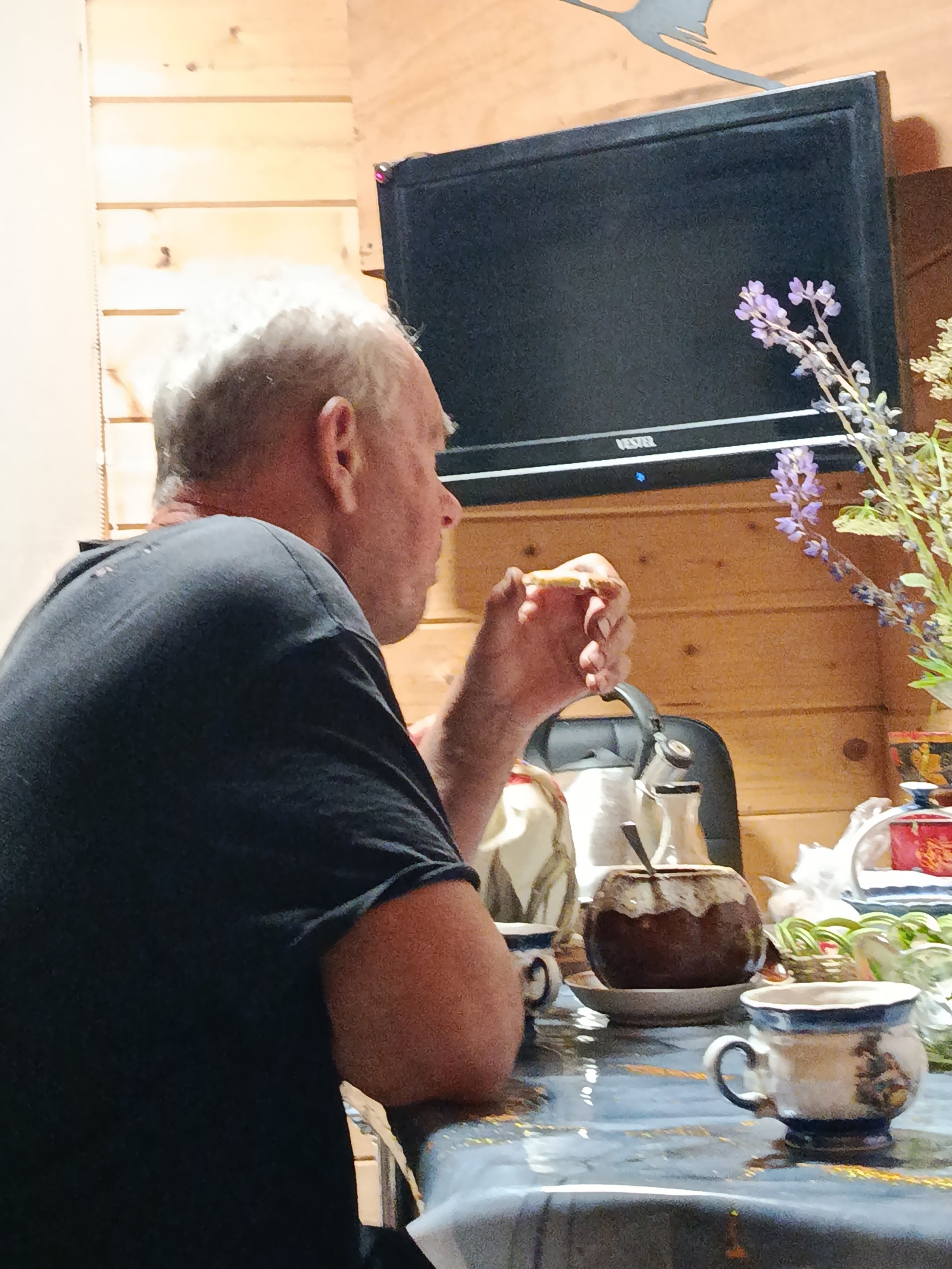 A medium shot captures a man sitting at a table, viewed from his left side. He has short, white hair and is wearing a dark t-shirt. His left hand is raised to his mouth, holding a piece of food. In front of him, on the table, are several items including a light-colored teapot, a brown ceramic pot with a spoon, and two ceramic mugs with blue and white designs. A bouquet of purple flowers is visible to the right of a black television screen mounted on a wooden paneled wall. The table surface is patterned with a dark blue and white design. The environment appears to be an indoor setting, possibly a rustic or cabin-like room due to the wooden walls.