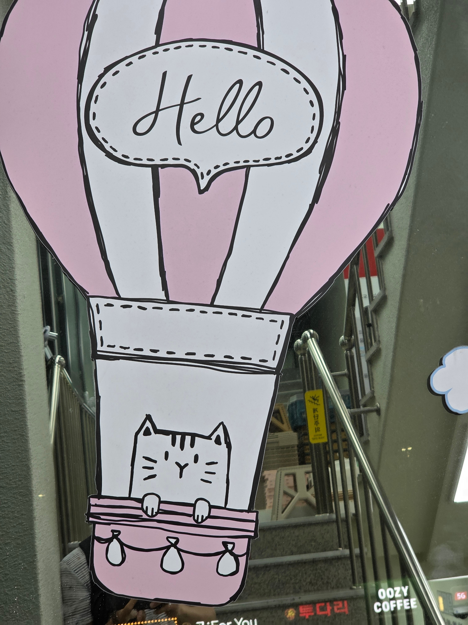 This image shows a decorative sticker on a glass surface, likely a storefront window, in Daejeon, South Korea. The sticker features a cartoon drawing of a pink and white hot air balloon. Inside a speech bubble on the balloon, the word "Hello" is written in cursive black lettering. Peeking out from the side of the balloon is a black and white cartoon cat with its paws resting on the balloon's edge. Attached to the balloon are several small, white balloons with black outlines. The background of the sticker is white, with a black outline and dashed stitch details adding to the cartoonish aesthetic. The reflection on the glass reveals an indoor setting with metal railings and what appears to be a display of items, possibly in a cafe or shop. Visible text in the reflection includes "OOZY COFFEE" and some Korean characters that read "투다리". The lighting suggests it is daytime. The overall mood is cheerful and inviting, conveyed through the playful illustration and the welcoming "Hello" message.