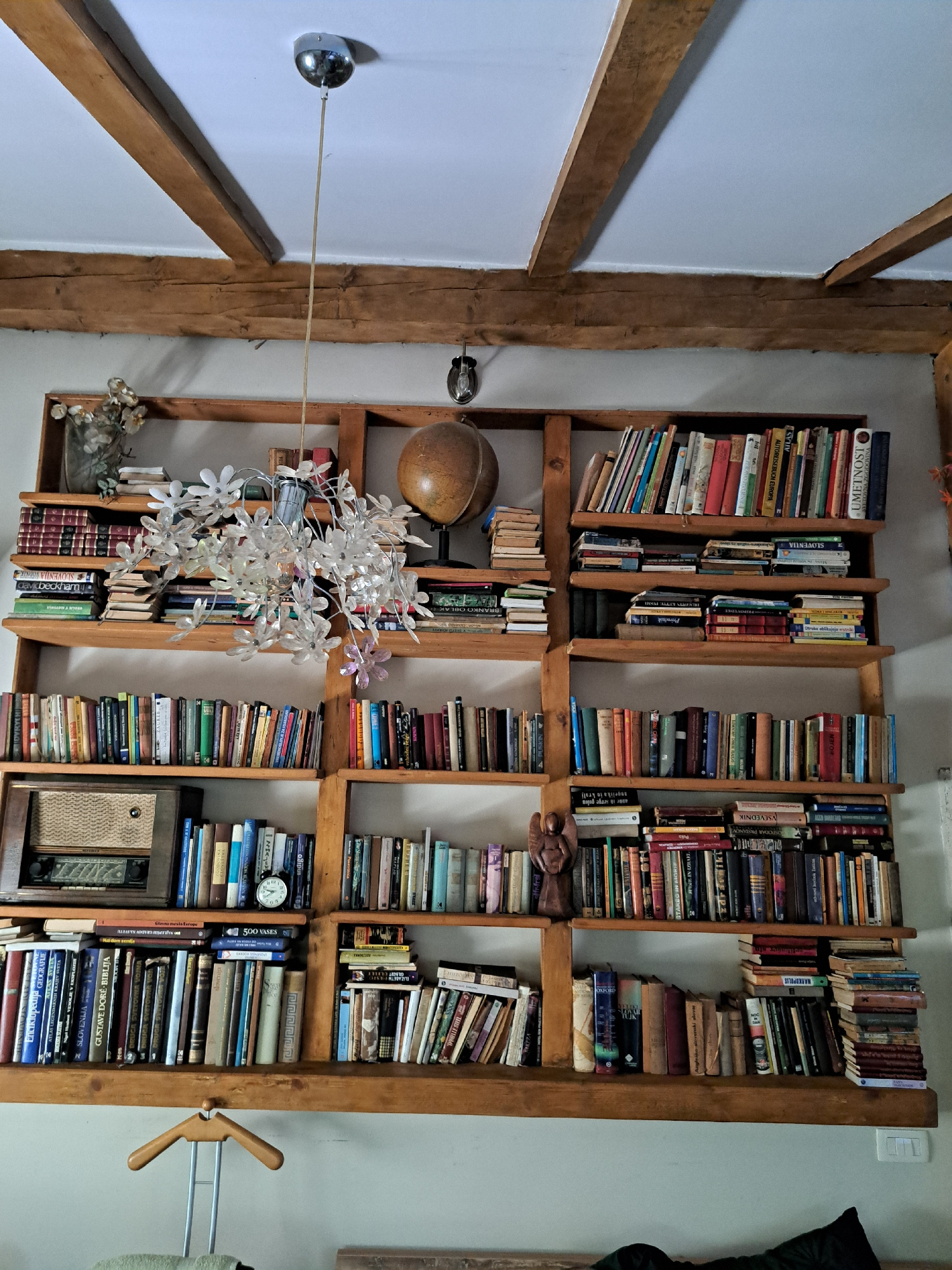 The image depicts a rustic interior scene featuring a large, floor-to-ceiling wooden bookshelf filled with books. The bookshelf is constructed from natural wood, with visible grain and some imperfections, suggesting an aged or handcrafted piece. It is divided into several horizontal shelves, densely packed with a variety of books, some appearing to be hardcovers and others paperbacks, with spines displaying different colors and titles.
On the top shelf, to the left, a glass vase with dried flowers and a stack of books are visible. A decorative chandelier with clear floral-shaped elements hangs in front of the center of the bookshelf. To the right of the chandelier, a globe on a stand is placed on a shelf.
Further down, the bookshelf houses a vintage radio on the left side of a lower shelf, accompanied by a small, round clock. Another section of the bookshelf features more books, including a small wooden figurine of an angel on one of the shelves. The overall environment suggests a cozy and scholarly atmosphere, possibly a living room or study. The lighting appears to be a mix of natural and artificial, casting some shadows. The walls are painted a light color, and wooden beams are visible on the ceiling, enhancing the rustic aesthetic. The image is taken from a slightly upward angle, emphasizing the height of the bookshelf.