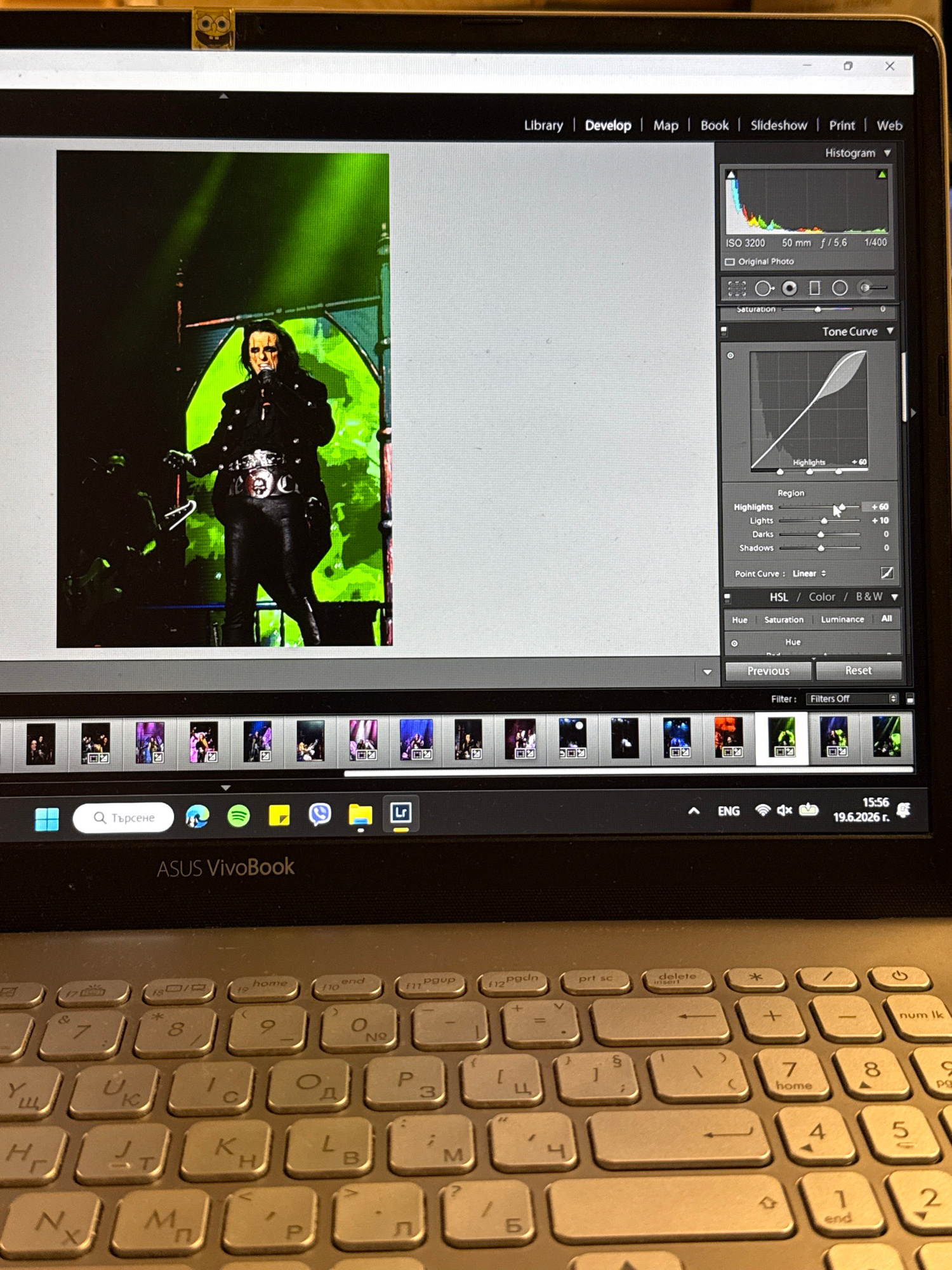 The image shows a laptop screen displaying Adobe Lightroom software. The main image on the screen is a photograph of a musician with white face paint and dark clothing, performing on stage with a bright green background. To the right of the image are various editing panels within Lightroom, including a histogram, tone curve, and sliders for adjusting highlights, lights, darks, and shadows. Below the main image is a row of thumbnail previews of other photos, likely from the same photo session. The bottom of the screen shows the Windows taskbar with application icons and system information, including the date "19.6.2026 r.". The laptop itself is an ASUS VivoBook, and a Spongebob Squarepants webcam cover is attached to the top bezel of the screen. The image appears to have been taken in Plovdiv, Bulgaria, given the date format and the Cyrillic characters on the keyboard.
