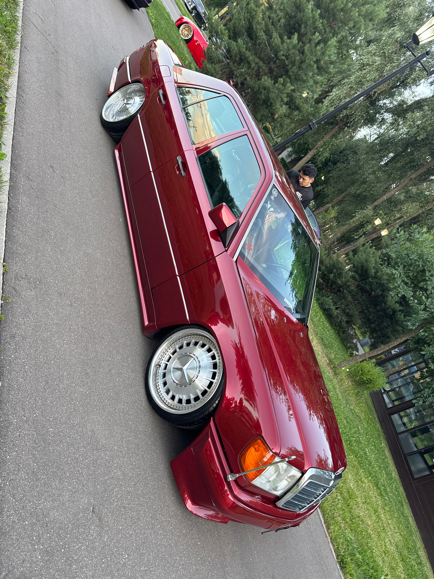 A low-angle, full-shot, exterior, vertical-format, eye-level, medium-shot photo captures a cherry red Mercedes-Benz W124 wagon on asphalt. The car is modified with a lowered suspension and aftermarket rims. The car's driver's side is closest to the camera, angled to the right. The vehicle is parked on a paved area that is bordered by a grassy area with trees and a building on the right. The car's license plate is not visible. A young man is leaning into the driver's side window. The scene is outdoors during the day, with the sun casting reflections on the car's body. The image appears to have been taken at an outdoor car meet in Moscow, Russia.