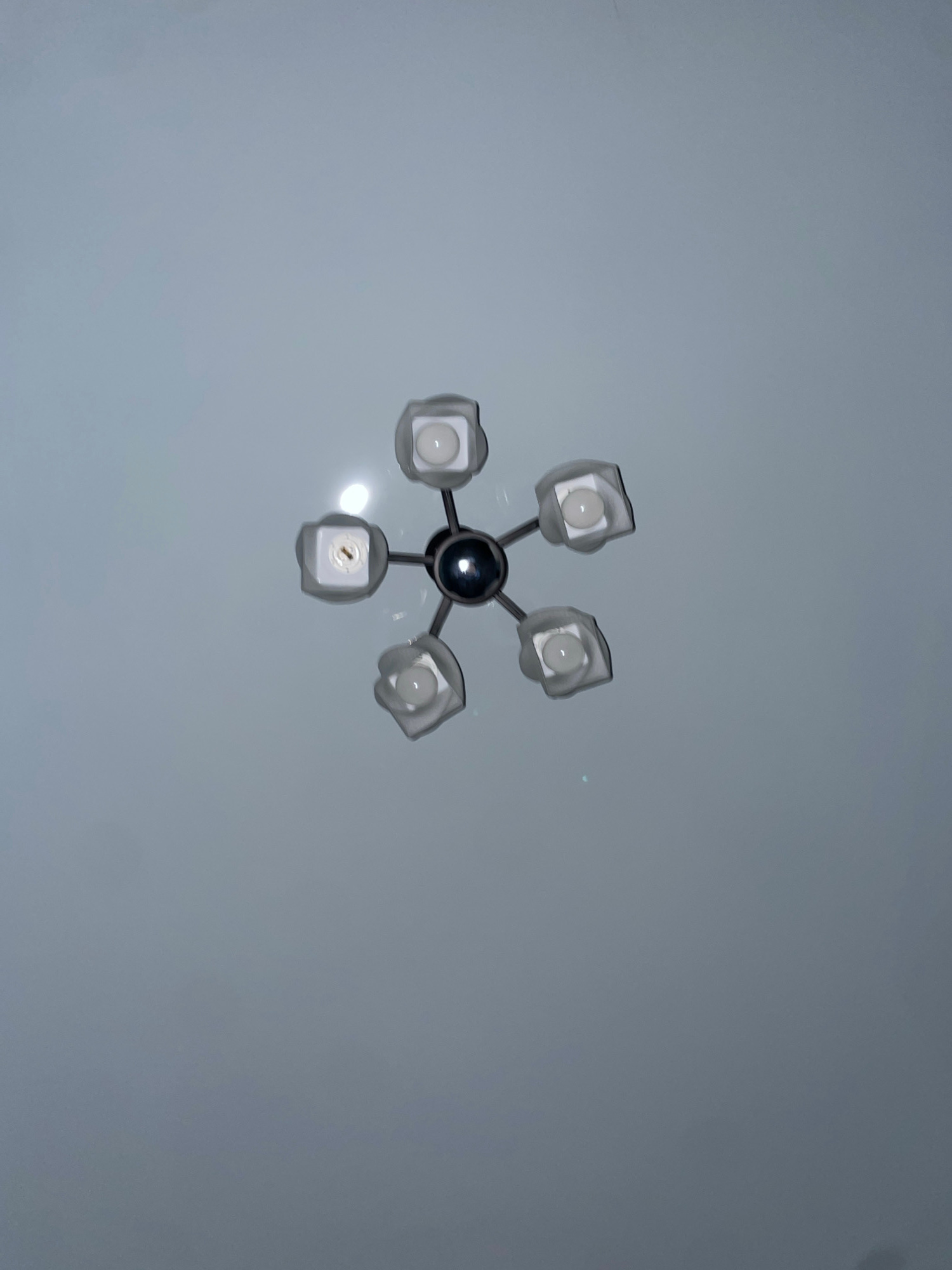 An overhead shot shows a ceiling light fixture against a plain, light-colored ceiling. The light fixture is suspended from the center and has a central spherical element with five arms extending outwards. Each arm terminates in a frosted, square-shaped light cover, and within each cover, a white bulb is visible. One of the bulbs appears to be a different type, with visible internal components. The overall impression is of a modern, somewhat abstract light design. The location is not discernible from the image.