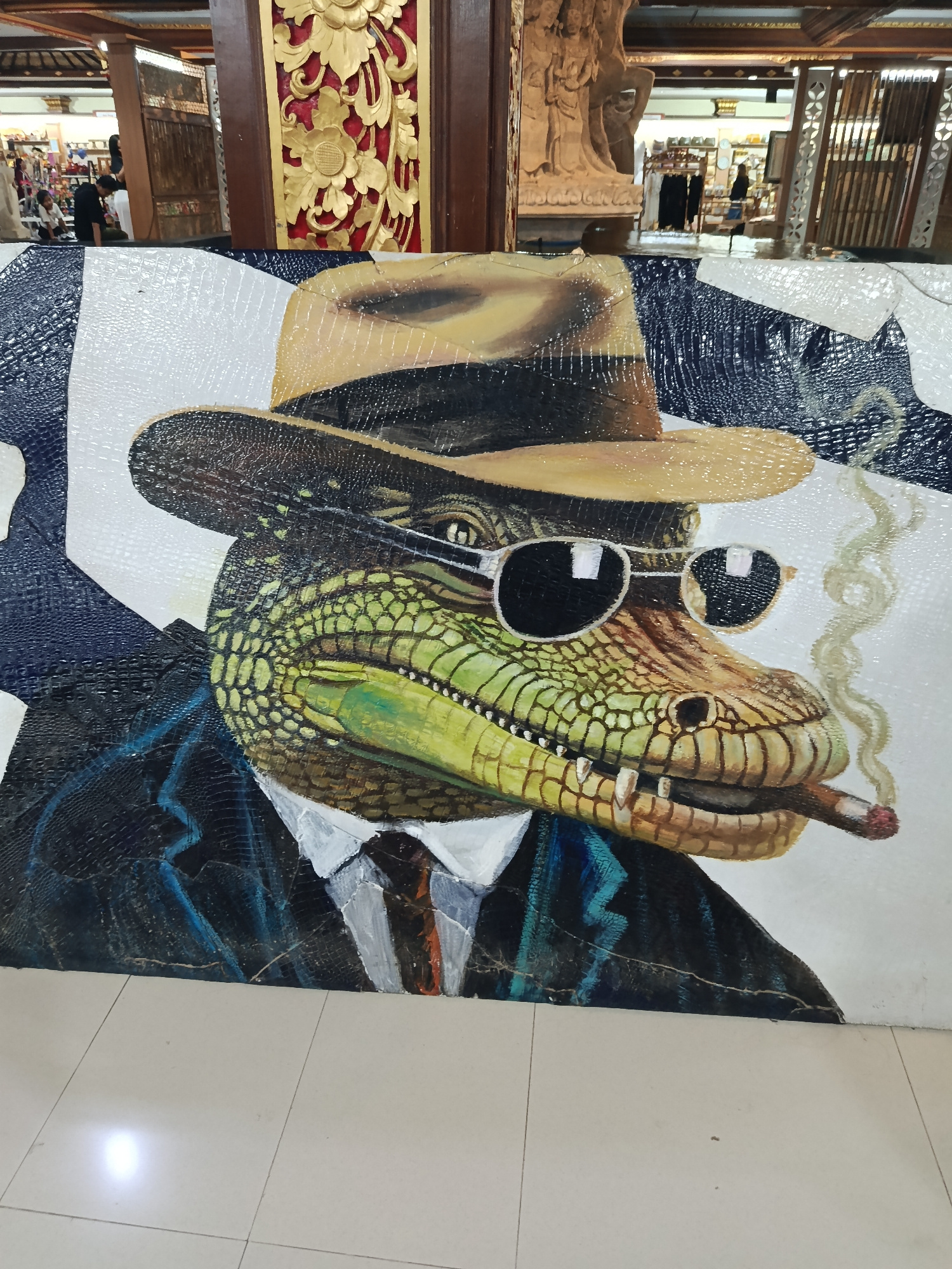 The image shows a large, detailed mosaic of an alligator wearing a fedora hat, sunglasses, a suit, and a tie, holding a lit cigar. The mosaic is mounted on a white background and appears to be a standalone art piece, not part of a larger scene. The alligator's skin texture is rendered in a variety of greens and browns, with yellow accents. The suit is dark blue or black with a lighter blue shirt and a striped tie. The hat is light brown, and the sunglasses have black lenses. Smoke is depicted rising from the cigar.
The background of the image is blurred but suggests an indoor setting. In the upper portion of the image, there is wooden architectural detailing, possibly part of a building's interior. Behind and to the left of the mosaic, there are glimpses of people and what appears to be a market or shop area with displays of goods. The floor in front of the mosaic is tiled with large, light-colored square tiles. The lighting suggests an indoor environment with some ambient light sources. The overall impression is of a piece of art displayed in a public indoor space, possibly a market, gallery, or shop.
