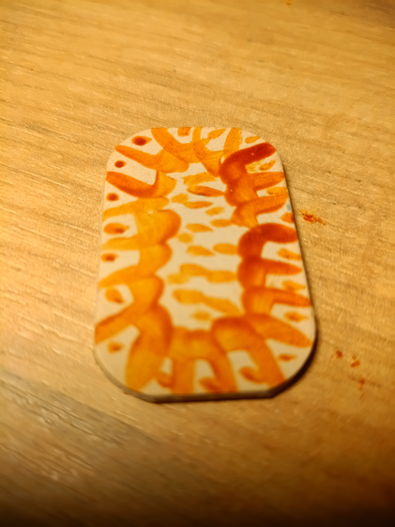 A close-up, eye-level, medium shot captures a small, light-colored, oval-shaped object with rounded corners, possibly a token or charm, resting on a wooden surface. The object is decorated with an abstract pattern of orange markings, resembling brushstrokes or splashes of paint. These orange marks form curved, repetitive shapes with some smaller dots scattered between them. The wooden surface appears to be a table or countertop, exhibiting a visible grain pattern and a light brown to golden hue. The lighting casts a soft glow on the scene, highlighting the textures of both the object and the wooden surface. There is no indication of the object's location in Irkutsk, Russia, or any environmental context beyond the wooden surface. No people, actions, or interactions are visible.