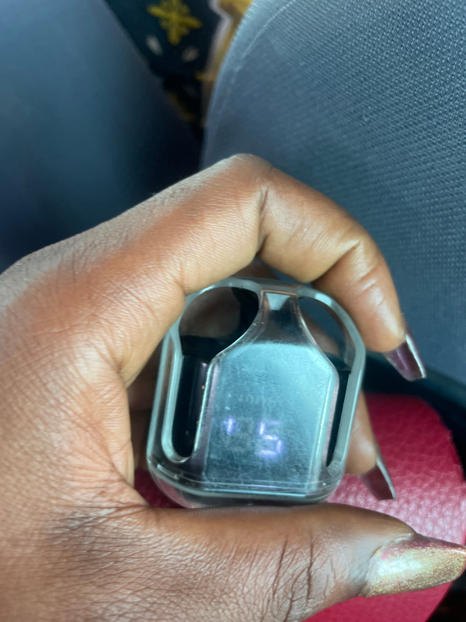 A close-up shot shows a person's hand holding a clear, rectangular electronic device with a digital display. The device is held in the palm, with the fingers wrapped around it. The digital display shows a numerical reading, appearing to be "10". The hand has dark skin and is adorned with manicured fingernails, some with a metallic gold glitter finish and others with a deep purple hue.
The background is out of focus, but appears to be an indoor setting. To the left, a textured, dark blue material is visible, possibly upholstery or fabric. To the right, a segment of a red, textured surface, resembling leather or a similar material, is present. The lighting suggests it is daytime, with artificial light contributing to the overall illumination. The context of Bauchi, Nigeria, is provided, but there are no specific visual cues within the image to confirm this location. The activity depicted is simply the holding of the electronic device, with no other discernible actions or events taking place. The image focuses on the hand and the device, with no visible text other than the numbers on the display.