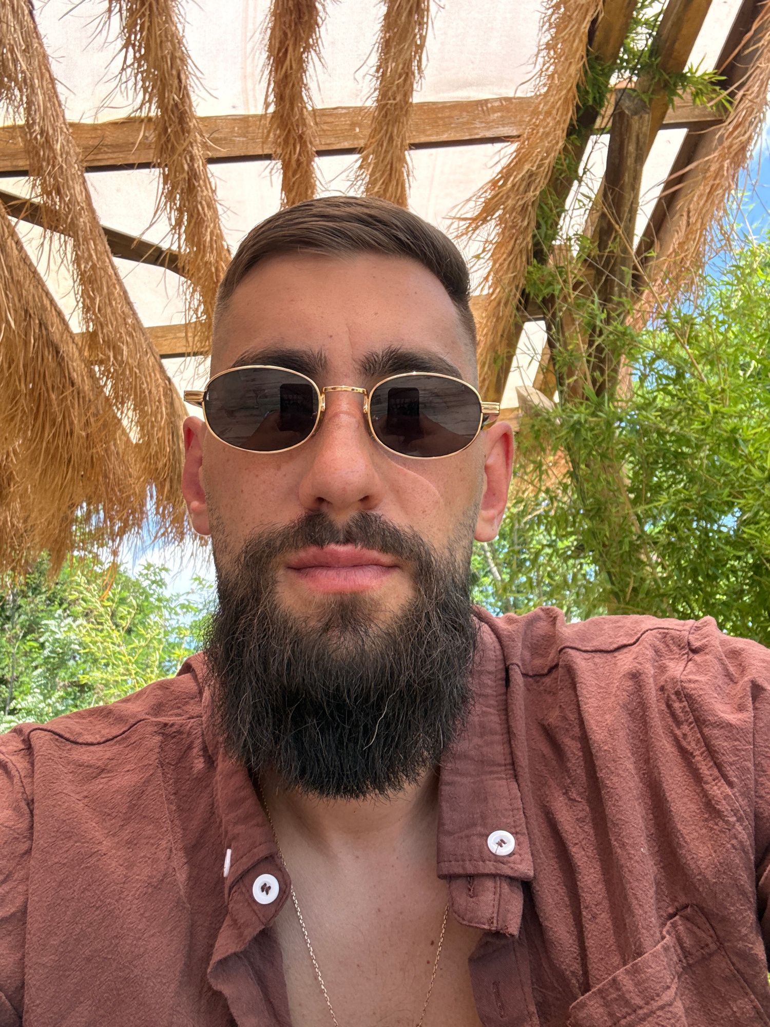 A close-up, vertically oriented, low-angle shot shows a man with a beard and sunglasses, wearing a brown button-up shirt. He is outdoors, with a thatched roof structure and green foliage visible in the background. The man's face is partially obscured by his sunglasses, but his mouth is visible. The scene suggests a warm, sunny day, likely in a tropical or Mediterranean location, given the vegetation and the straw-like roofing. The lighting indicates it is daytime. There is no visible text in the image. The man appears relaxed. The overall impression is of a casual, outdoor leisure setting.