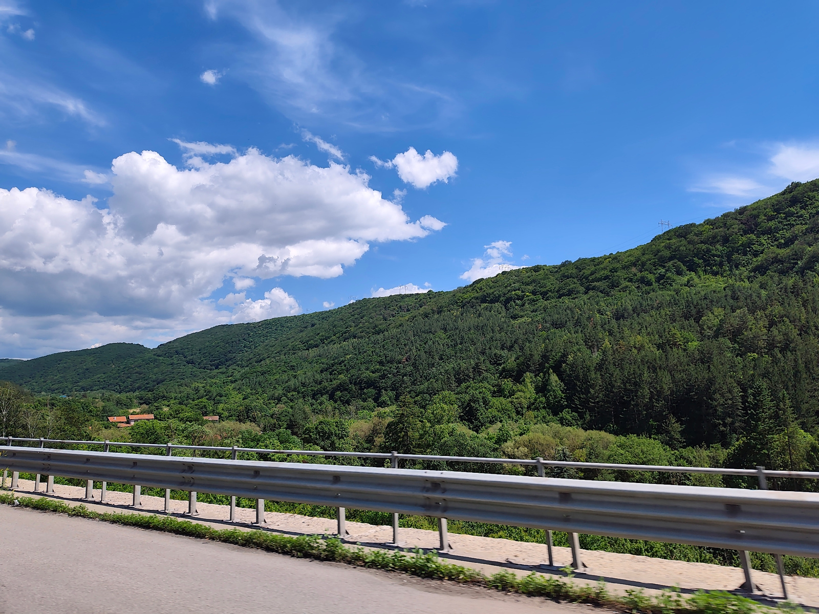 The image shows a scenic view of a green, forested mountainside under a blue sky with scattered white clouds. In the foreground, a metal guardrail runs along the edge of a paved road, which is visible in the lower part of the image. The road appears to be curving. Beyond the guardrail, the lush green vegetation of the mountain slopes is prominent, with a dense forest covering the terrain. In the middle ground, several houses with red tile roofs are nestled among the trees. The overall impression is one of a natural, rural landscape. The location is specified as the city of Potop, Bulgaria. There are no people visible in the image, and the scene depicts a static environmental view.