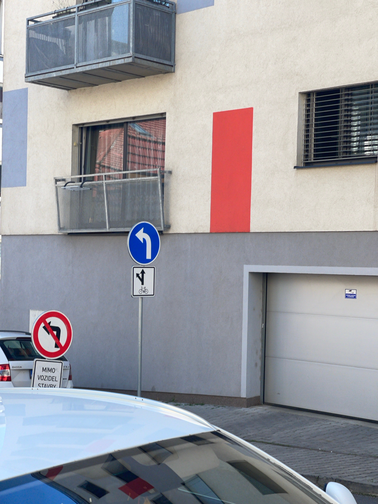 The image depicts a modern building with a light-colored facade, featuring a prominent red vertical stripe and balconies with metal railings. A blue circular traffic sign with a white left-pointing arrow is mounted on a pole, accompanied by a rectangular sign below it showing a left-turning arrow and a bicycle symbol. To the left, another circular traffic sign with a red border prohibits left turns, with a white background and a black arrow indicating a forbidden left turn. Below this sign, a white rectangular sign displays Czech text: "MIMO VOZIDEL STAVBY". A white Skoda car is partially visible on the left, and the roof of a white car is in the foreground, reflecting the building. A closed garage door is on the right side of the image. The scene is set on a paved walkway. This image was taken in Pilsen, Czechia.