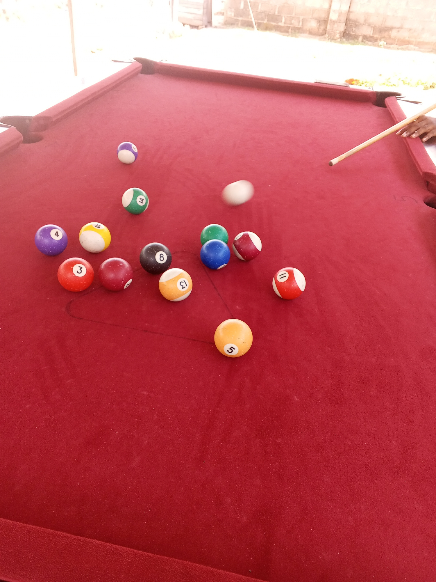 This image depicts a pool table with several billiard balls scattered on its red felt surface. A pool cue is positioned on the table, held by a person's hands, indicating an ongoing game or practice. The table is outdoors, with a brick wall and a wooden structure visible in the background, suggesting a location in Lokoja, Nigeria. The lighting suggests it is daytime. The billiard balls have various numbers and colors, characteristic of a standard set. Some balls are clustered together, while others are spread across the table. One ball appears to be in motion, possibly struck by the cue. The overall scene captures a moment of leisure activity in an outdoor setting.