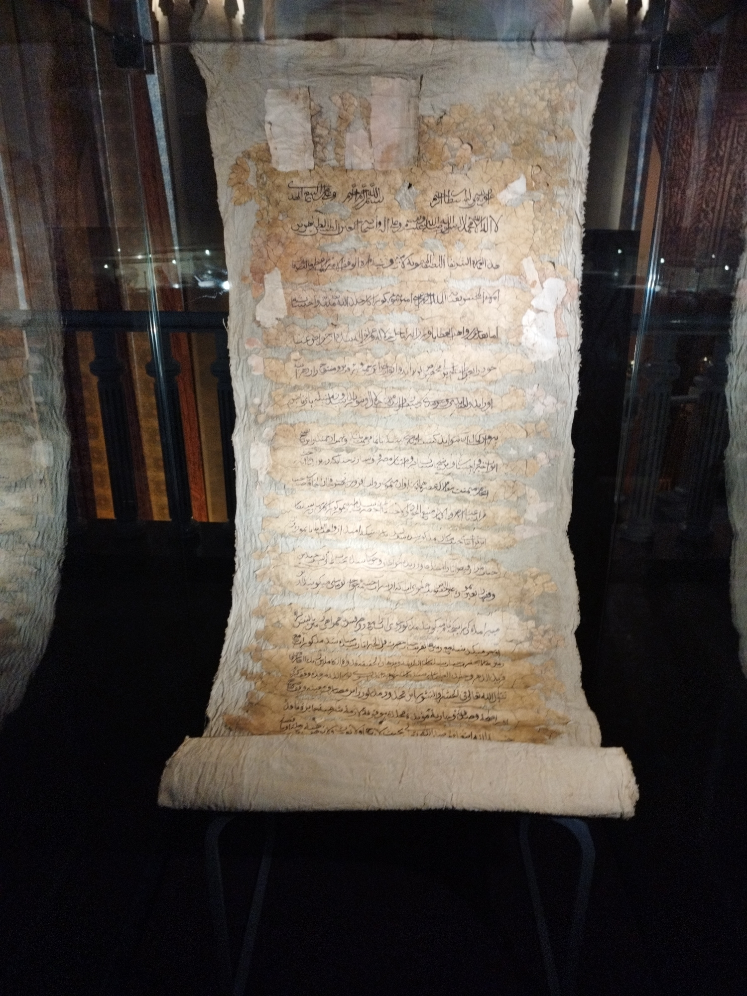 In a dimly lit, museum-like setting, a large, ancient scroll is displayed vertically, partially unrolled and mounted on a dark stand. The scroll is made of what appears to be parchment or aged paper, with a rough, uneven texture and visible signs of wear and tear, including tears, creases, and discoloration. The text on the scroll is written in Arabic script, in dark ink, and is arranged in multiple lines. The script is a historical form, suggesting significant age.
The scroll is framed by glass, and the background is dark, with the hints of red and brown striped curtains or wall coverings visible through the glass on the left and right sides. The lighting is directed from above, casting shadows and highlighting the texture of the scroll. The bottom of the scroll is rolled and rests on a black metal stand. The overall impression is of an artifact preserved for historical and cultural display. There are no people visible in the image. The environment appears to be an indoor exhibition space, likely a museum or gallery.