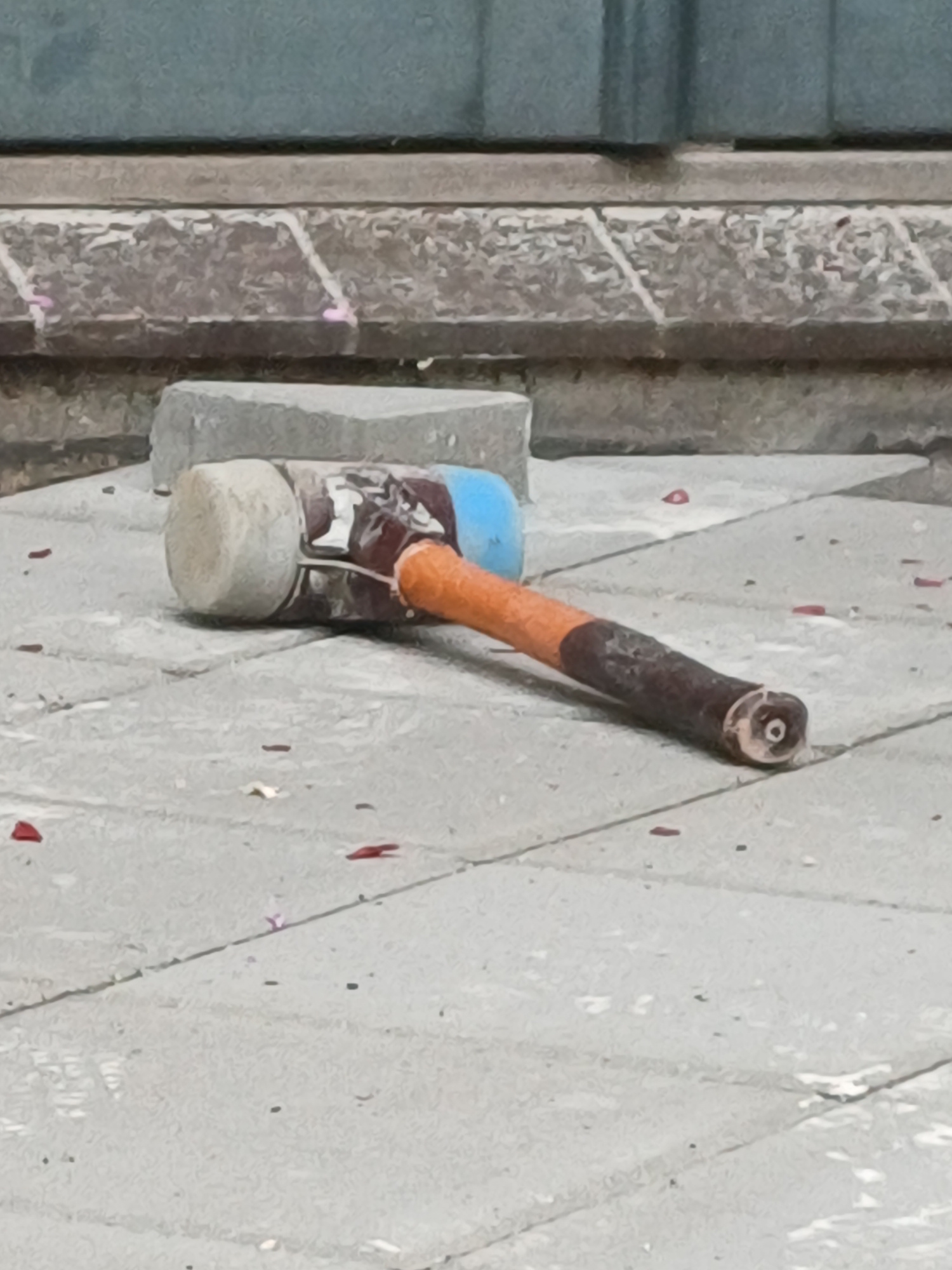A rubber mallet lies on a paved outdoor surface. The mallet has a white rubber head, a blue and brown body, an orange grip, and a dark brown handle. The mallet is positioned diagonally on the ground. The ground appears to be made of interlocking pavers. There are small red and pink pieces of debris scattered on the ground around the mallet, potentially confetti or flower petals. In the background, there is a stone wall with a textured surface and a darker, smooth surface above it. The image does not contain any identifiable landmarks or specific elements that would allow for pinpointing a location within Copenhagen, Denmark.