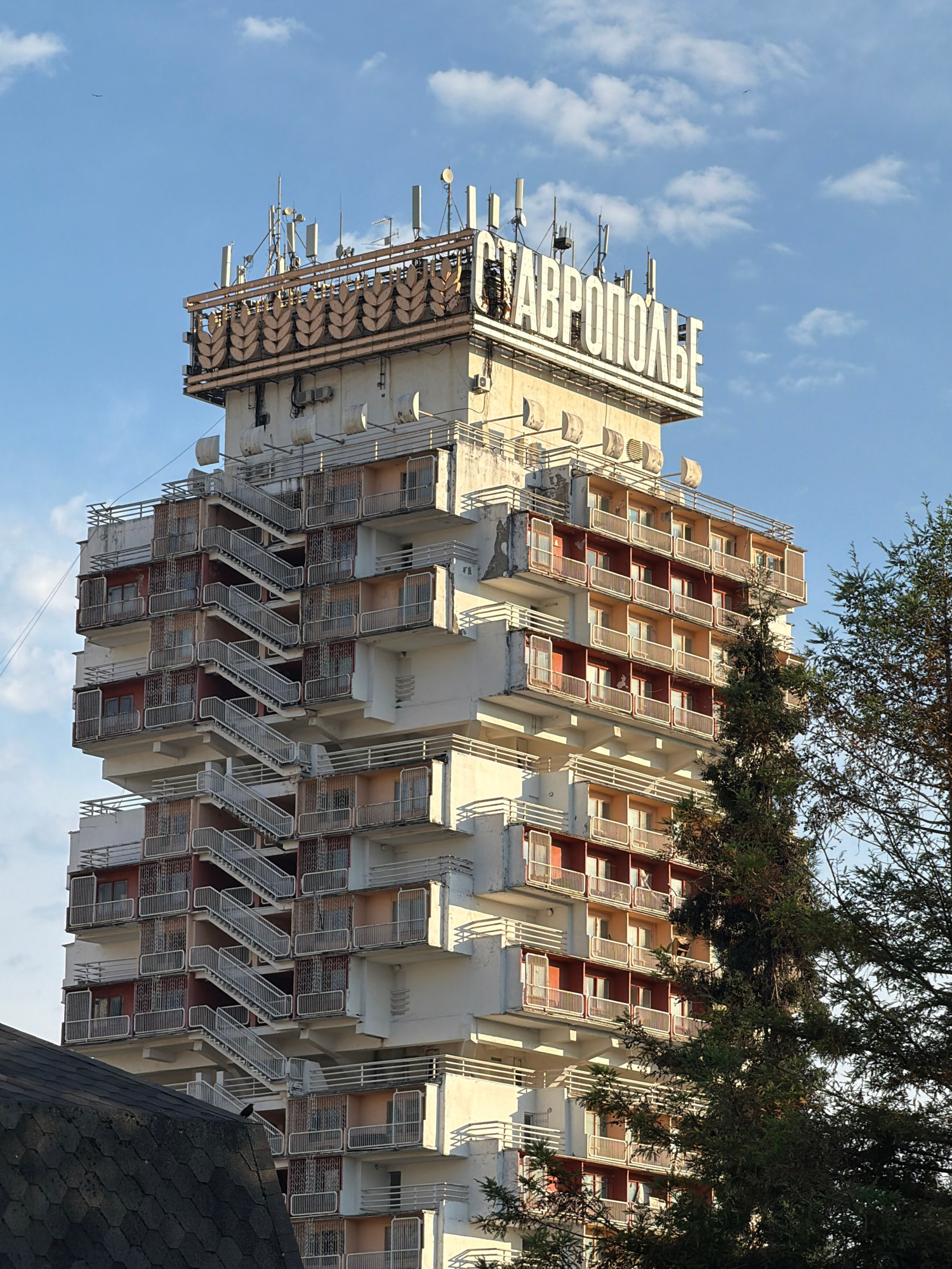 A large, multi-story building with a distinctive tiered facade featuring numerous balconies and external staircases is shown in Sochi, Russia. The building is topped with a sign that reads "СТАВРОПОЛЬЕ" in large, white lettering. The building is surrounded by trees and the sky is partly cloudy. The architectural style suggests a Soviet-era construction, possibly a hotel or residential complex.