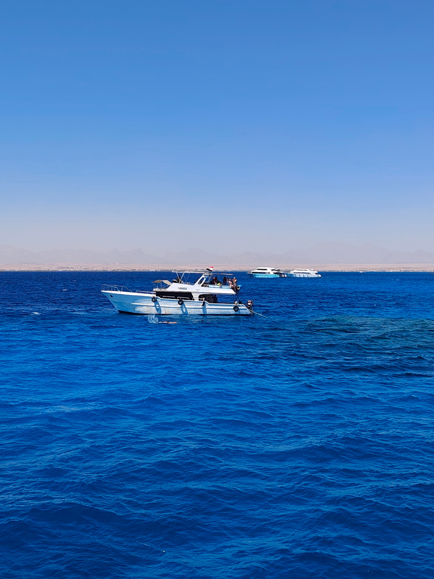 This image depicts a clear, bright day at sea in Hurghada, Egypt, with a wide expanse of deep blue water dominating the foreground and midground. The water exhibits a rippled texture, suggesting gentle waves. In the midground, a white motor yacht is anchored, with several people visible on its deck. The yacht has a dark blue stripe along its hull and a canopy over part of its upper deck. Further in the distance, another white boat with a blue section is visible. The background reveals a hazy, distant coastline with mountains. The sky is a uniform, light blue, devoid of clouds. The overall scene conveys a tranquil, aquatic environment, characteristic of coastal resorts like Hurghada.