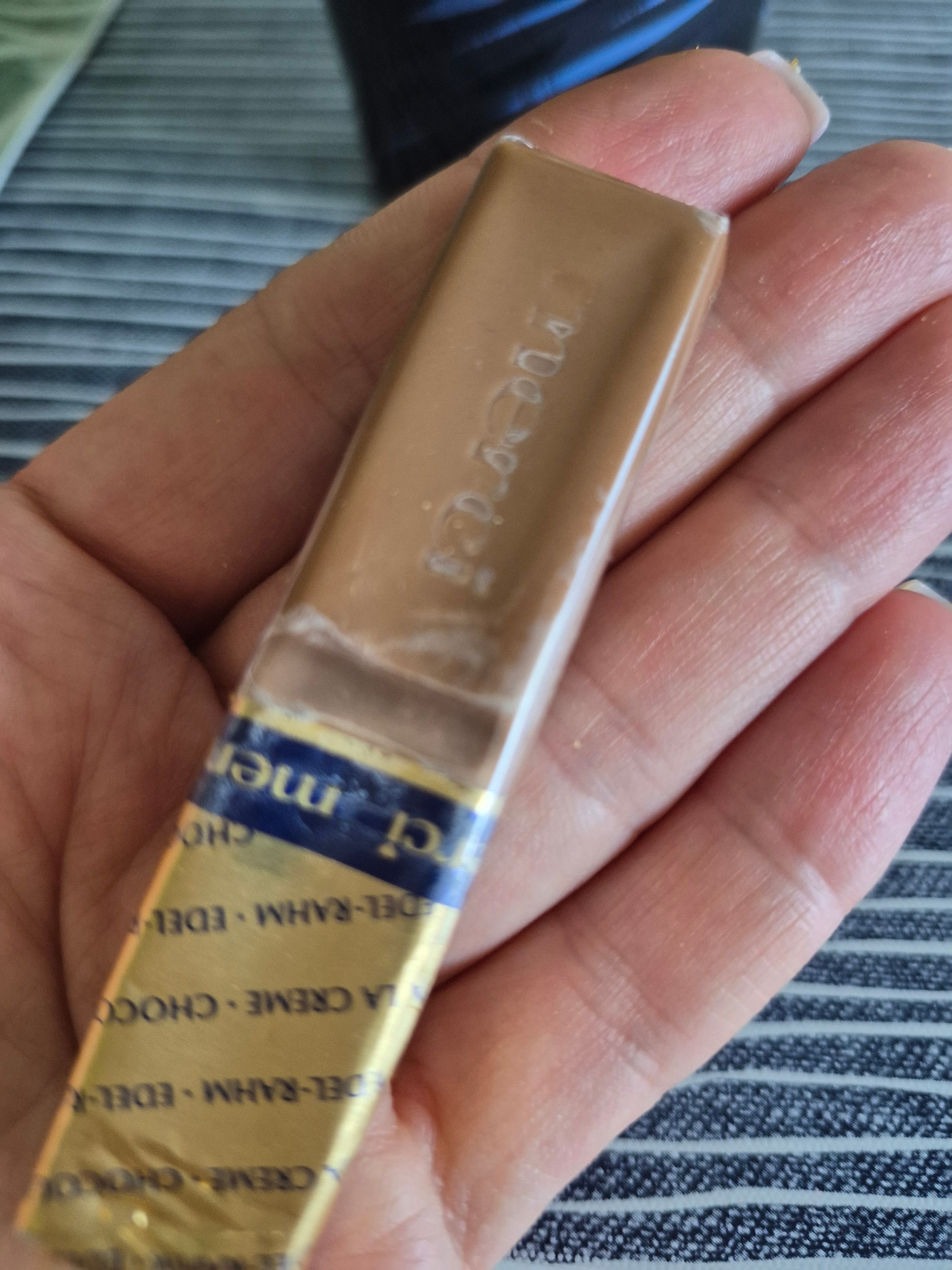 A person is holding a rectangular chocolate bar with gold and blue foil wrapper. The wrapper has some text that appears to be in German, including "Merci" and "Edel-Rahm Schokolade." The chocolate bar itself is light brown and has some text embossed on it, which is difficult to read due to the foil. The background is a striped fabric, possibly a tablecloth or placemat, in shades of blue and gray. The lighting is natural and appears to be from above. The scene is close-up, focusing on the hand and the chocolate bar.