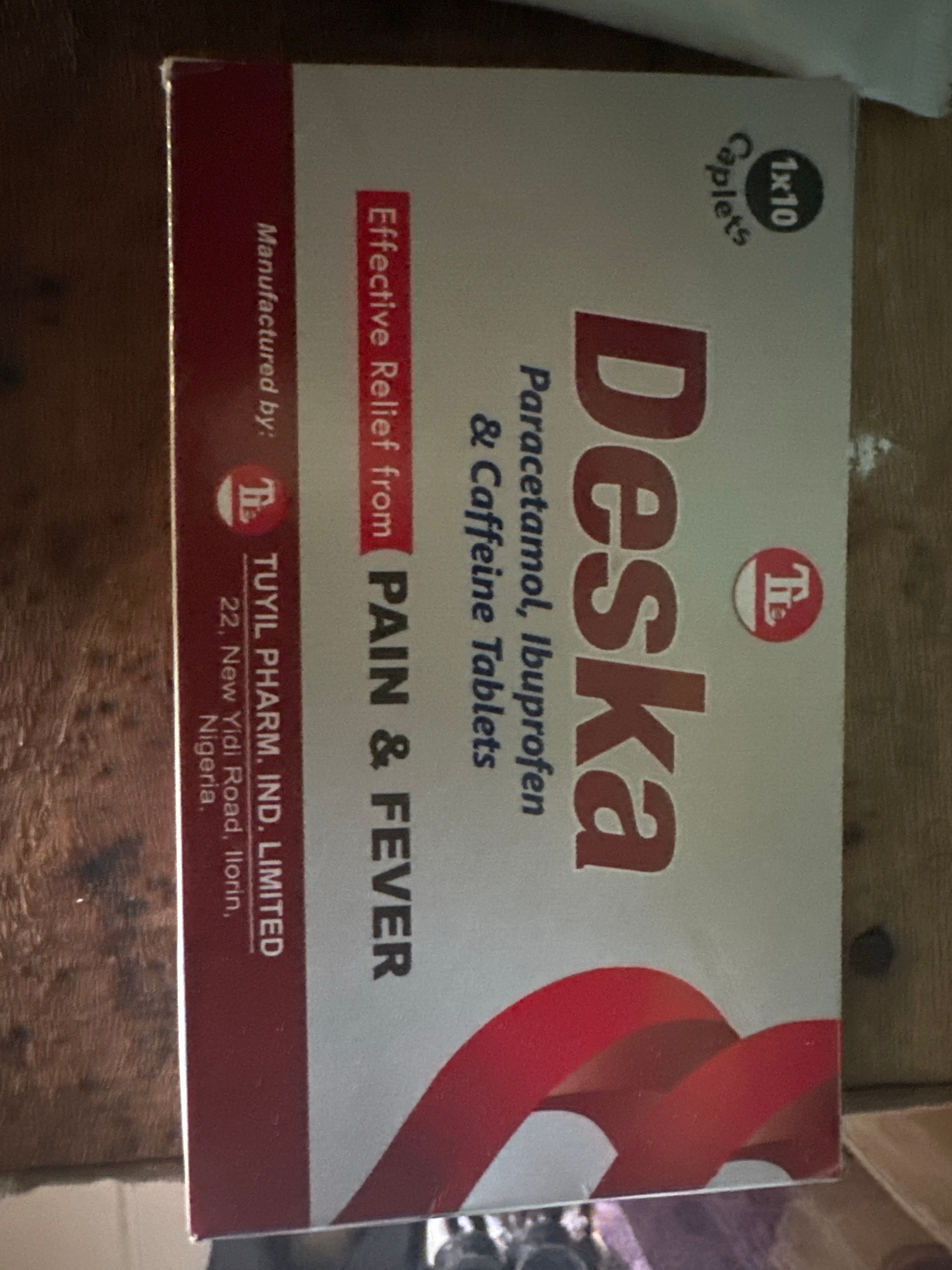 The image shows a vertically oriented pharmaceutical box for "Deska" tablets. The box is primarily white with a red side panel and a red curved design on the front. Text on the box indicates it is manufactured by "TUYIL PHARM. IND. LIMTED" located at "22, New Yidi Road, Ilorin, Nigeria." It also states "Effective Relief from PAIN & FEVER" and lists the contents as "Paracetamol, Ibuprofen & Caffeine Tablets." A small circular emblem with "1x10 Caplets" is visible in the upper right corner. The box is positioned on a wooden surface. The city of Jauro Yinu is not mentioned on the packaging, but the manufacturing address is in Ilorin, Nigeria.