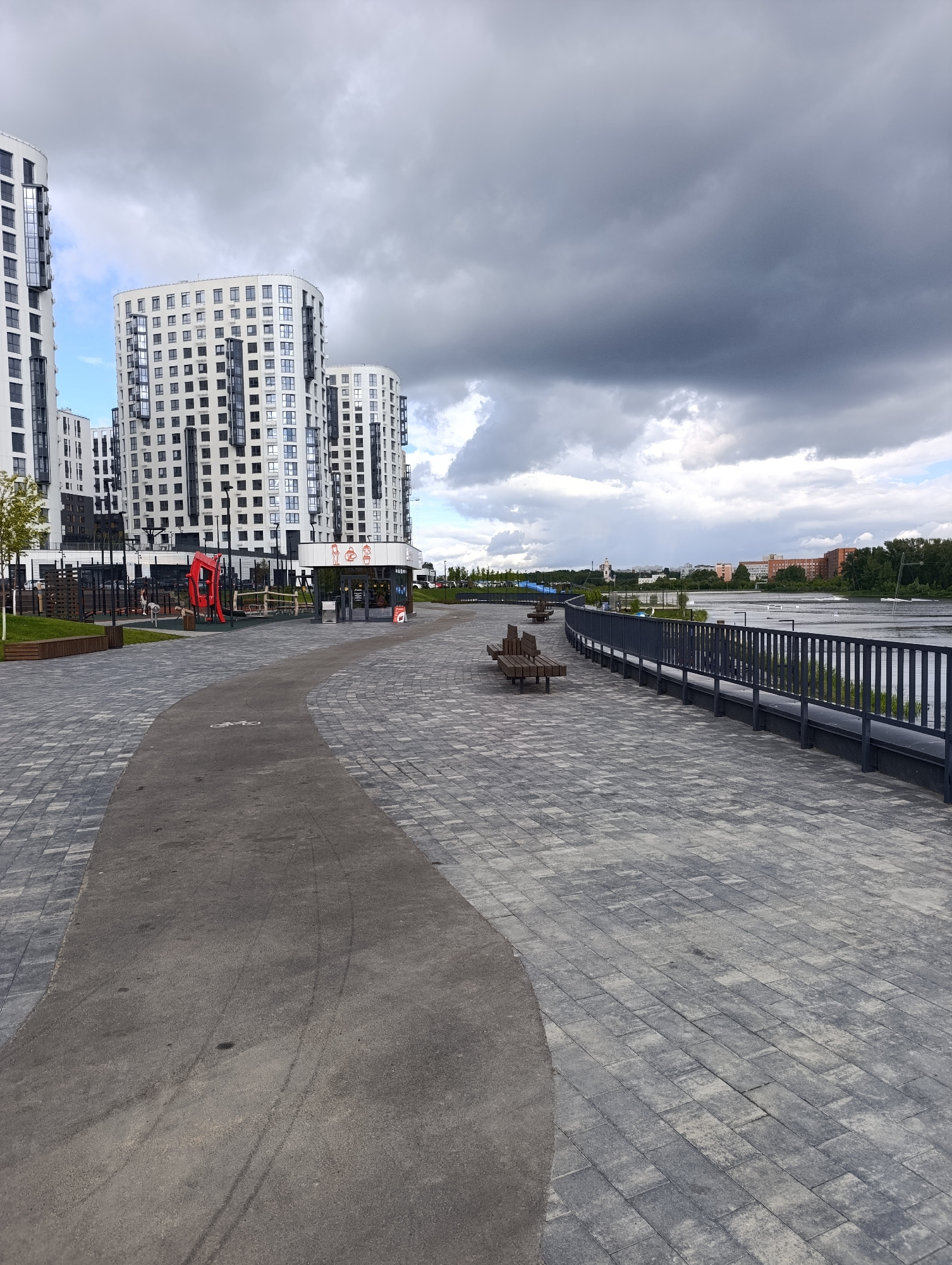 This is a horizontal, eye-level, full shot of a modern, paved walkway next to a body of water in Penza, Russia. The walkway is constructed from interlocking grey pavers and has a dark-colored asphalt path that curves through it, marked with a bicycle symbol. Along the right side of the walkway, a dark metal railing separates the path from the water. A few wooden benches are placed along the walkway, offering seating.
In the background, on the left side of the image, stand several tall, modern apartment buildings with white facades and numerous windows. Adjacent to these buildings is a children's playground with red and yellow equipment. A small, white building with a red roof, possibly a cafe or kiosk, is also visible.
The body of water in the background appears to be a river or lake, with distant buildings and trees lining its far bank. The sky is filled with dramatic, dark grey clouds, suggesting an overcast or impending storm. Despite the weather, the scene feels serene and peaceful, with no people visible. The time of day is difficult to ascertain due to the cloudy conditions, but it appears to be daytime. No discernible text is visible in the image.