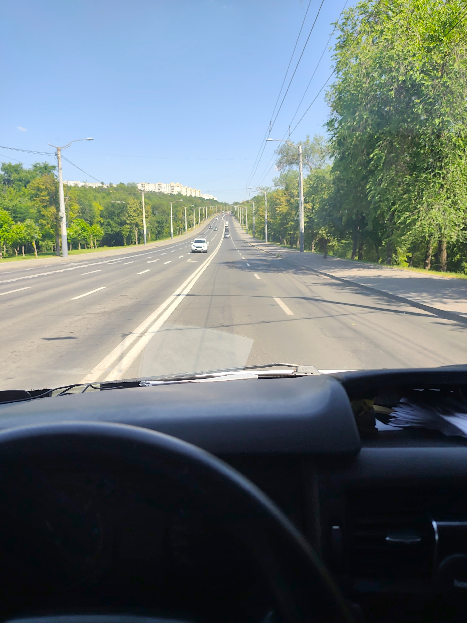 This media file depicts a view from the driver's seat of a vehicle, looking out at a multi-lane road in Chișinău, Moldova. The scene is set on a bright, sunny day, suggesting it's daytime. The road stretches into the distance, lined with trees and streetlights on both sides. In the background, apartment buildings can be seen on a hillside, providing a clue to the urban environment. Several cars are visible on the road, indicating moderate traffic. The foreground shows parts of the vehicle's interior, including the dashboard and the top of the steering wheel, suggesting the perspective is from someone driving. The overall atmosphere is neutral, portraying a typical scene of daily life on a road in a city. There is no visible text in the image.