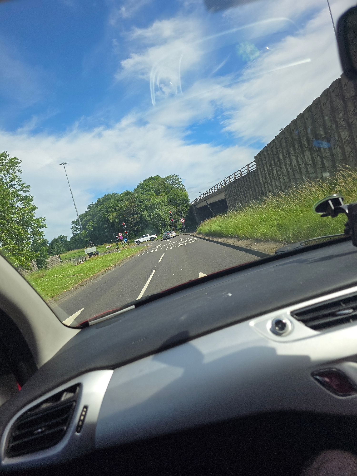 The image depicts a view from inside a car, looking forward through the windshield. The car is on a road with two lanes in the direction of travel. There are traffic lights and road signs visible. In the distance, trees line the road, and a bridge crosses overhead. The sky is blue with some white clouds. The dashboard of the car is visible in the foreground. The location is the A66 Eastbound, near the Stockton Road interchange in Middlesbrough, United Kingdom.