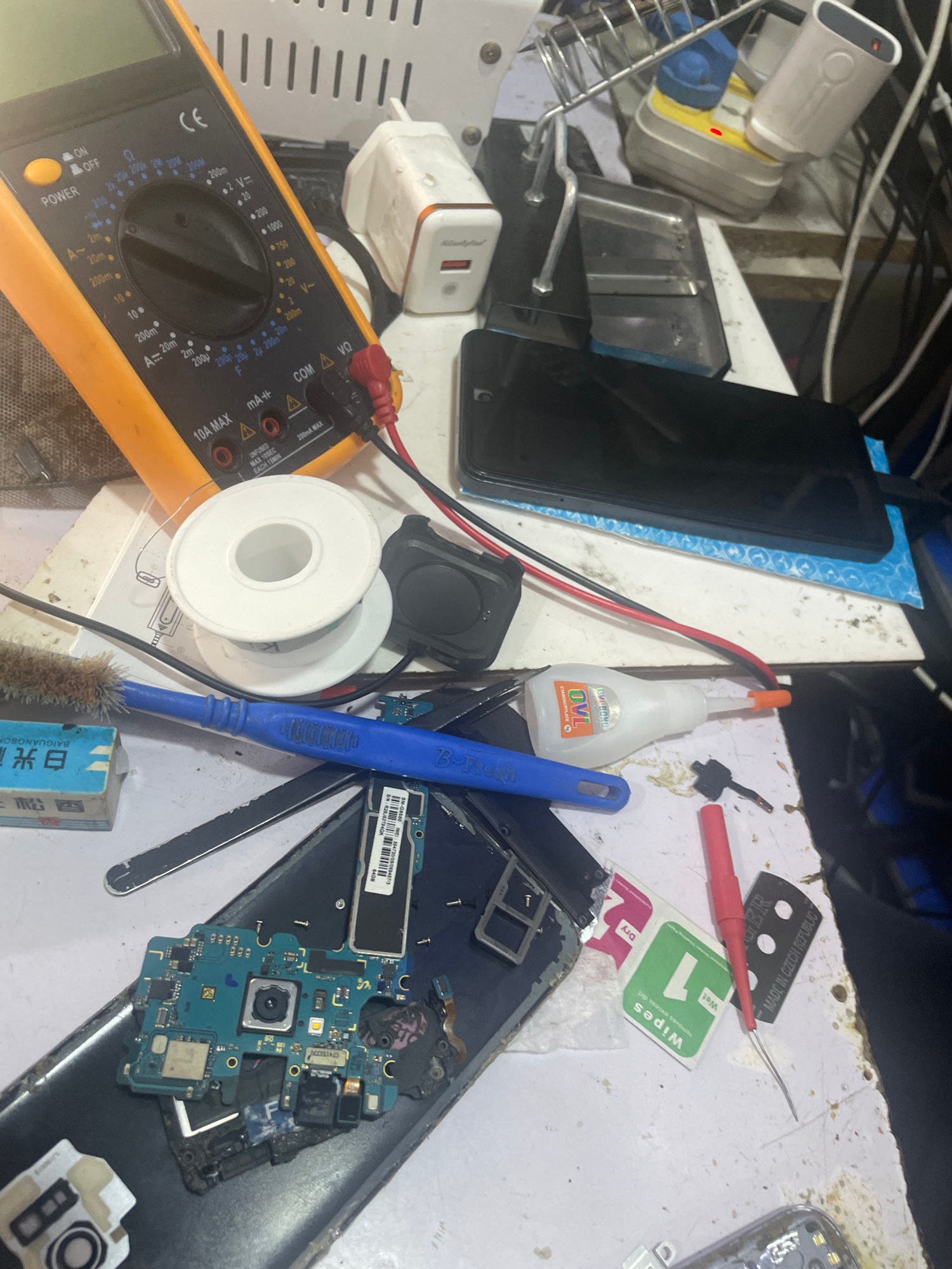 The image displays a cluttered workbench with various electronic components and tools, suggesting a repair or assembly process. In the foreground, a bright orange multimeter with a digital display is prominent, alongside spools of wire and a soldering iron. Several disassembled parts of what appear to be mobile phones are scattered across the surface, including circuit boards, screens, and casings. There are also charging adapters, a bottle of adhesive, and cleaning wipes, all indicative of electronic device maintenance. The overall scene is that of a busy, functional workspace, likely a small repair shop or a personal electronics tinkering area. The lighting suggests an indoor environment, possibly during daylight hours given the overall brightness. The visible text includes brand names and technical specifications on the components and tools, such as "OVL", "CE", and IMEI numbers on a phone circuit board. The location context of Jalingo, Nigeria, implies this is a typical scene of electronic repair services available in that region. No people are visible in the image.