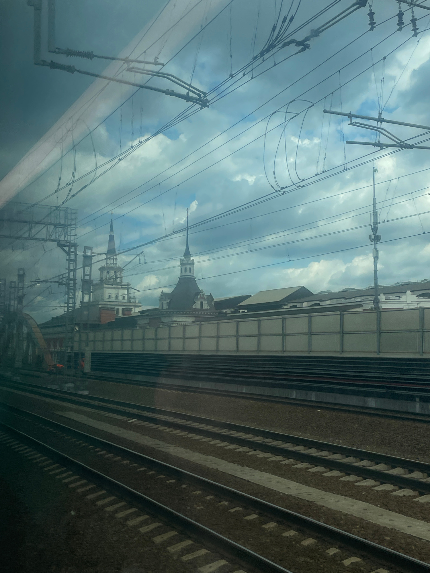 This image captures a view from a train window, looking out at a cloudy sky and a train station. Multiple sets of train tracks are visible in the foreground, leading towards a long, grey building with a patterned facade. Above the building, two distinctive white towers with dark spires rise into the sky. These are likely part of a historical building, possibly a train station or administrative building. A complex network of overhead power lines for electric trains crisscrosses the sky, obscuring parts of the view. The overall scene suggests an urban environment, likely a major transportation hub in Moscow, Russia, given the architectural style and the presence of extensive railway infrastructure. There are no people visible in the frame. The lighting indicates it is daytime, but the clouds create a diffused, somewhat somber atmosphere.