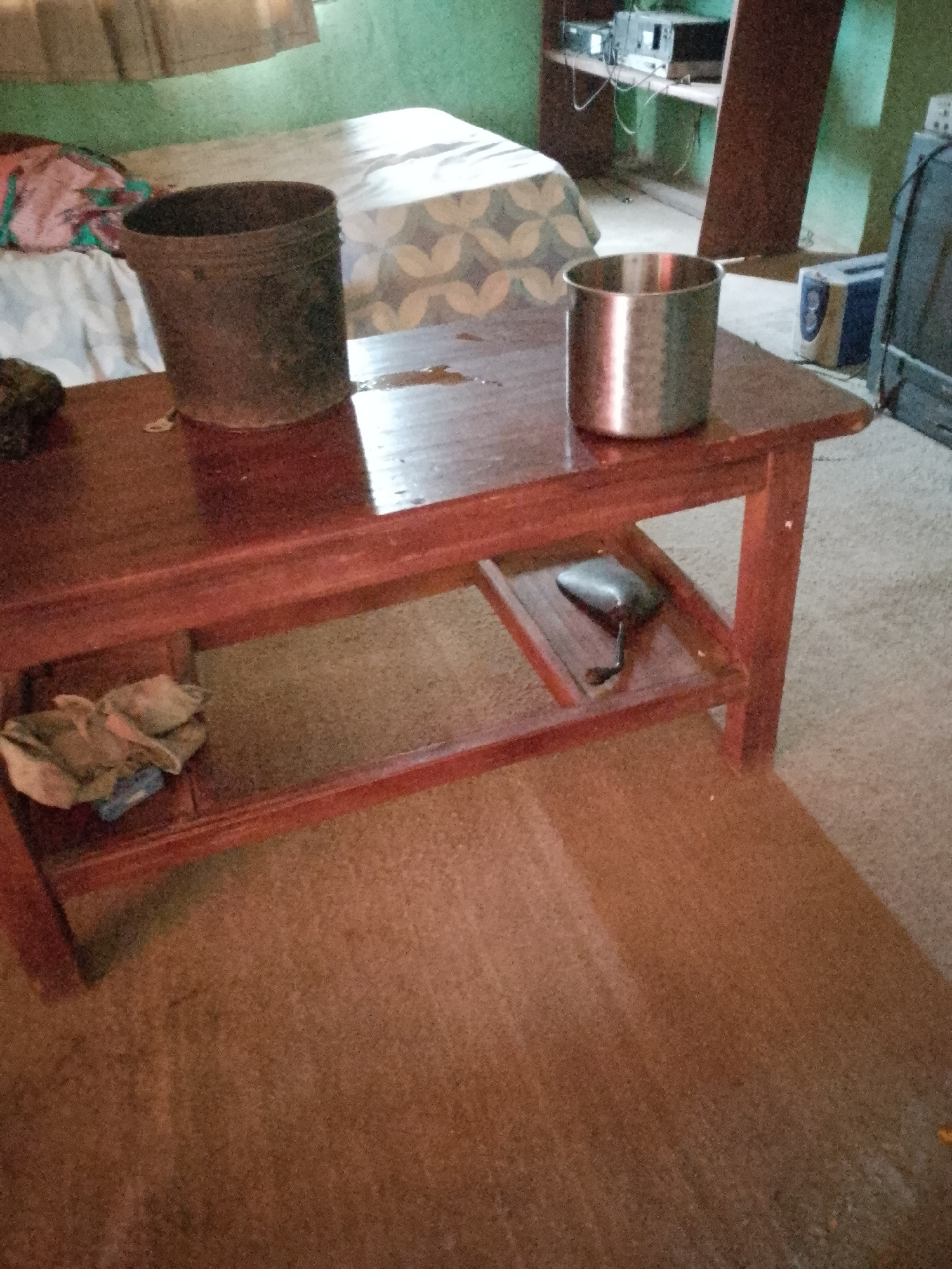 A wooden coffee table with a lower shelf sits on a carpeted floor in an indoor setting. Two metal containers are on the tabletop. The one on the left is a dark, rust-colored bucket, and the one on the right is a shiny, silver-colored cup. A wet spot is visible on the tabletop between the two containers. On the lower shelf, a dark, irregularly shaped object with a cord is placed to the right of center. On the left side of the lower shelf, a bundle of light-colored fabric is present. The background shows a bed with a patterned comforter, a wall painted green, and some electronics on shelves.