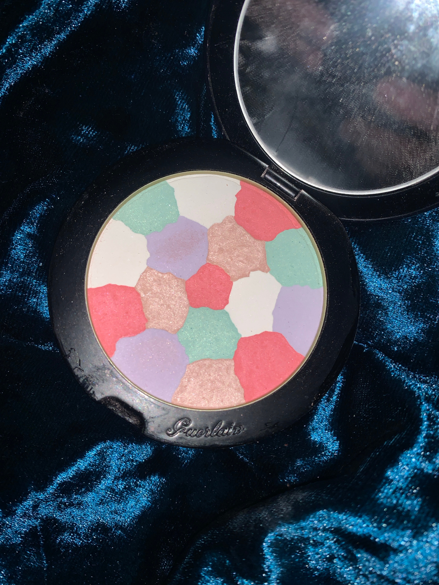 A close-up, top-down shot shows an open cosmetic compact with a multi-colored powder inside. The powder is divided into irregular, pebble-like shapes in shades of white, pink, light purple, mint green, and a peachy shimmer. The compact itself is black with a reflective lid, partially open, revealing a mirror. The Guerlain logo is embossed on the lower edge of the compact. The compact rests on a dark blue, velvety fabric with a rich, textured surface that catches the light, creating bright, sparkling highlights. The background is dominated by this shimmering blue fabric.