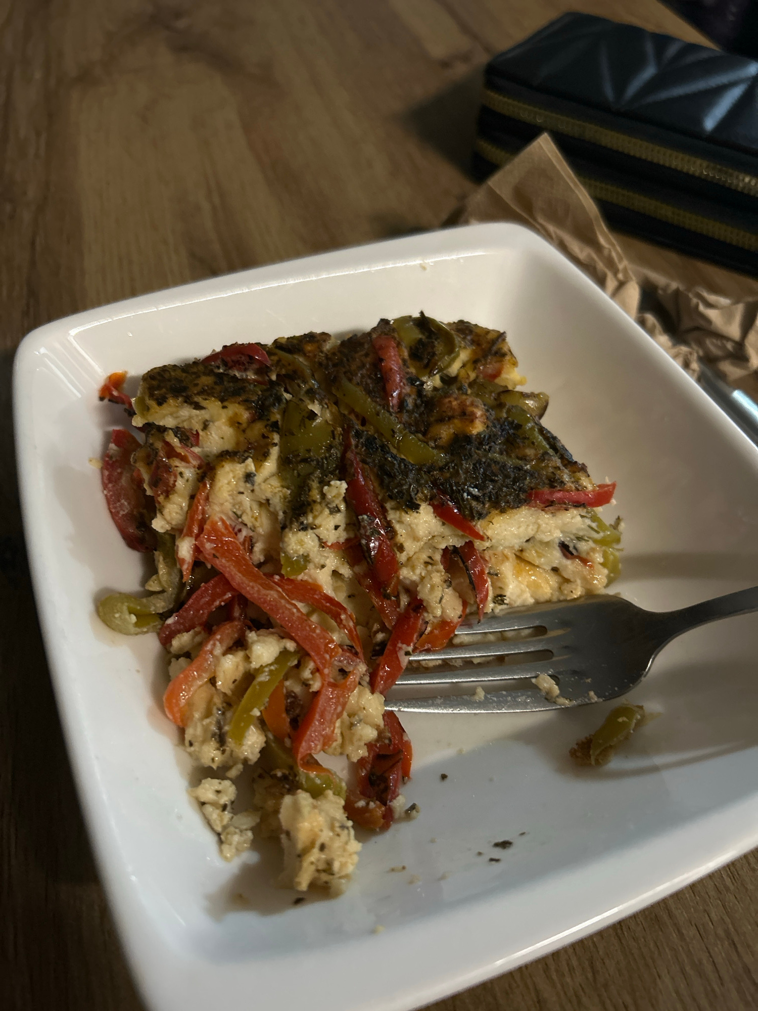 A white rectangular plate sits on a wooden table. A fork rests on the right side of the plate, partially in a slice of food. The food is layered, with a dark, herb-crusted top, a creamy white middle layer, and a bottom layer containing sliced red and green bell peppers and possibly egg. The plate is mostly filled with the food, with a small portion of the white plate visible on the right side. In the background, a dark object with a quilted pattern and gold trim, possibly a wallet or purse, rests on a brown paper bag. The wooden table has a visible grain pattern.