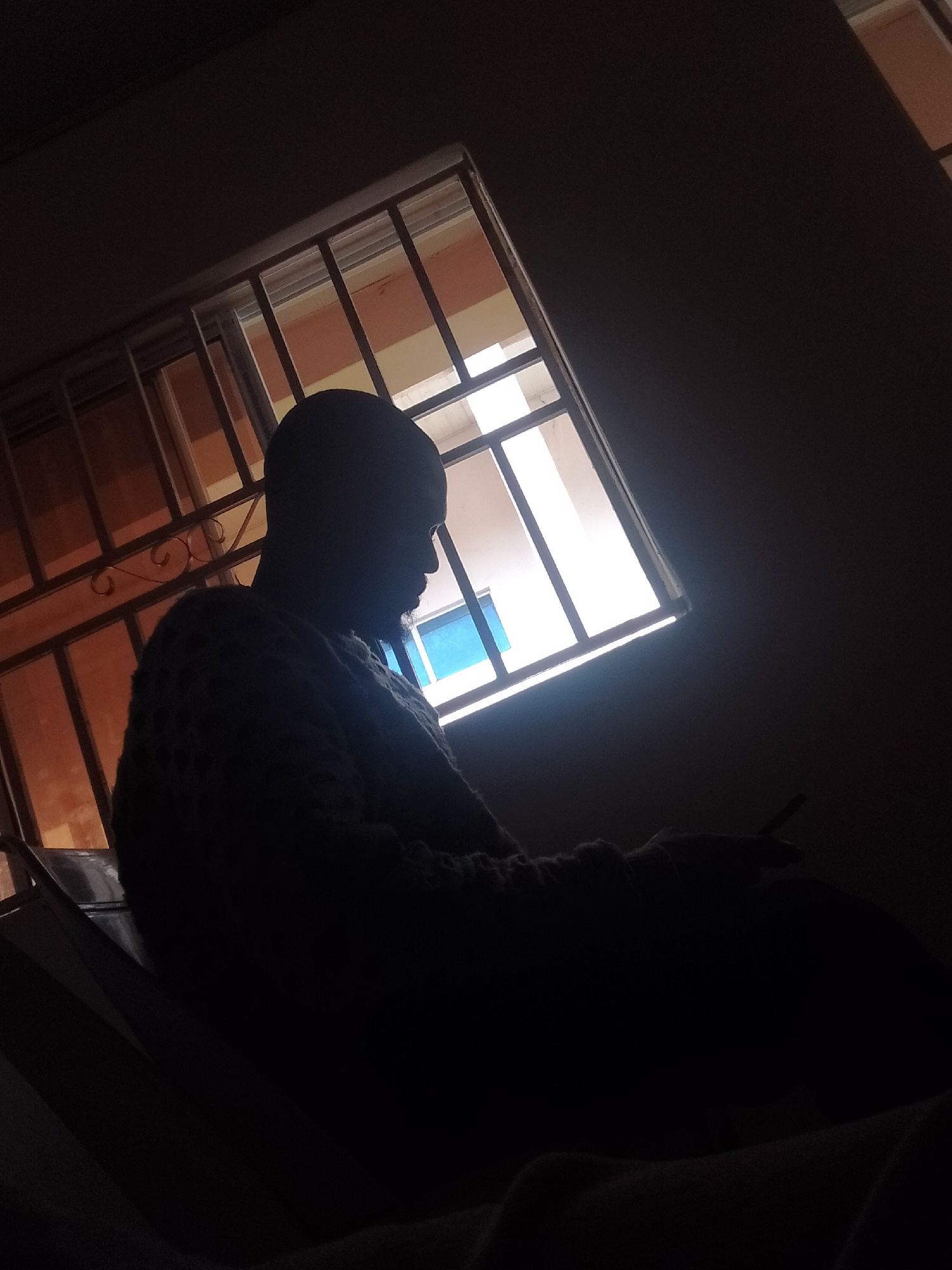 A silhouetted person, with a patterned sweater, sits facing away from the camera, looking out a barred window. The person's head is tilted down. Their right hand is raised, holding a cigarette. The window is partially illuminated by an external light source, casting a diagonal beam across the scene. The environment appears to be indoors, with a dark ceiling and wall visible.