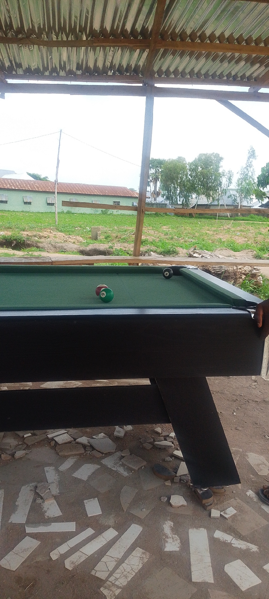 An eye-level, medium shot captures a pool table situated outdoors under a corrugated metal roof supported by wooden beams. The pool table's surface is green felt, and three pool balls are visible: one solid green ball with the number 9, one red and white striped ball, and one black ball. To the left of the table is a building with a weathered, reddish-brown roof and light green walls. Beyond the building, there are trees and a cloudy sky. The ground in the foreground is a mixture of dirt and broken white tiles. A person's hand is visible on the right edge of the frame, resting on the pool table.