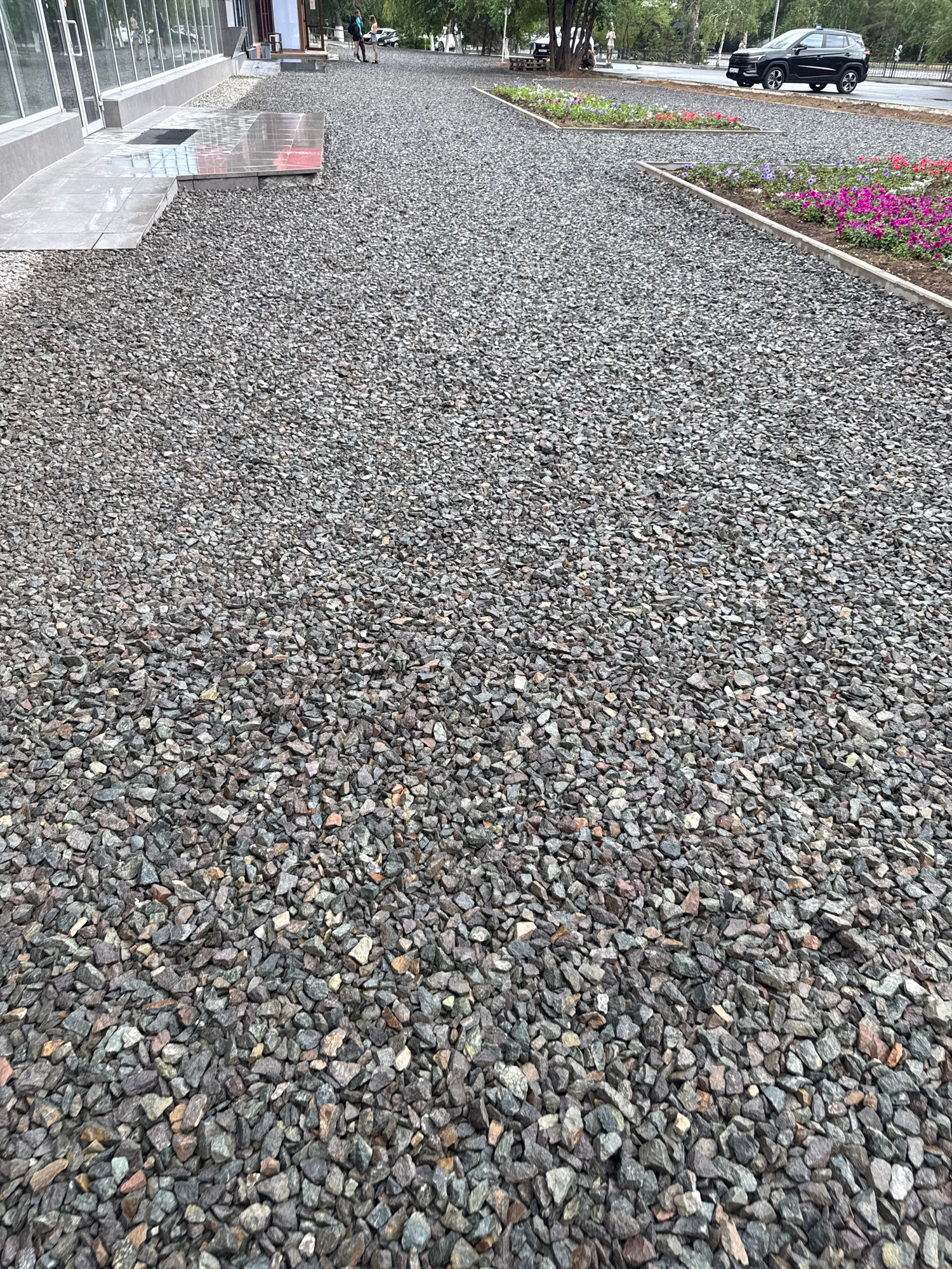 A wide shot of a gravel pathway leading towards a modern building with large glass windows, possibly a shop or office, in Pavlodar, Kazakhstan. The pathway is covered in dark grey and brown gravel. In the background, several people are visible walking. Further down the path, a black SUV is parked on the side of the road, and colorful flowers are planted in raised garden beds. The environment suggests an urban setting with a mix of commercial and natural elements.