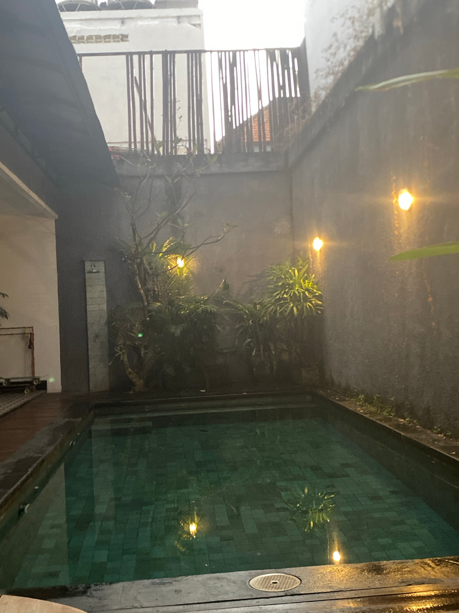 In North Kuta, Indonesia, a courtyard features a rectangular swimming pool with dark tiled flooring and reflective green water. The pool is surrounded by wooden decking. To the left of the pool, a tall, narrow structure resembling an outdoor shower stands against a grey wall. Lush green plants and a tree with visible branches are situated behind the shower. Several small, warm-toned lights are attached to the grey wall on the right side of the courtyard, illuminating the area. In the background, a white building with a slatted wooden fence on its upper level is visible, with what appear to be water tanks on its roof. The scene is dimly lit, suggesting it is dusk or nighttime.