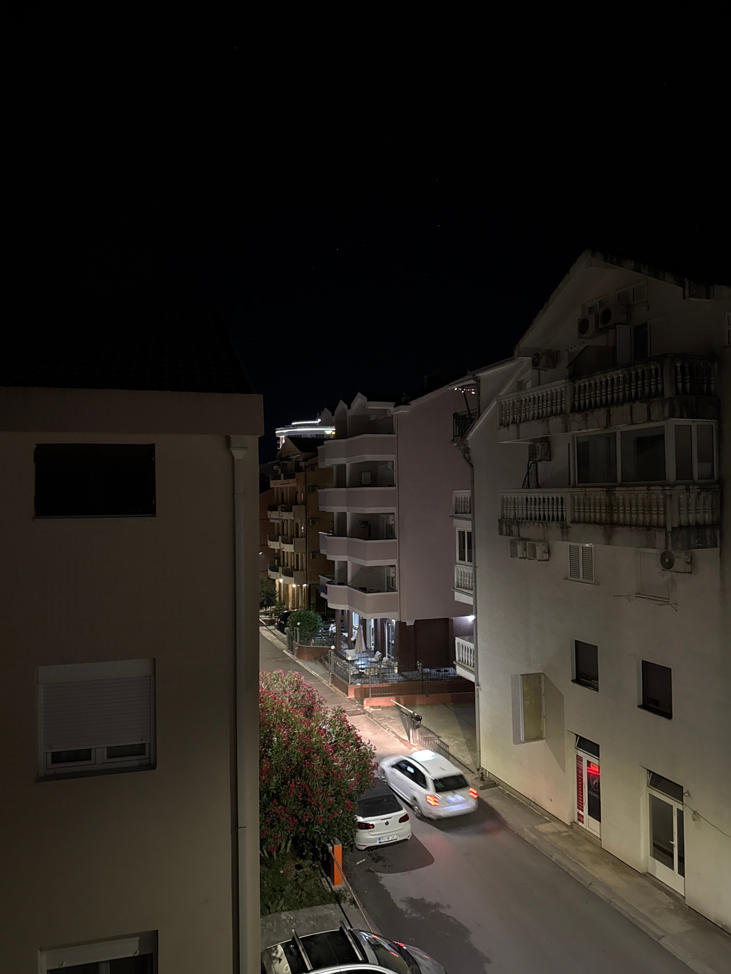 This is a nighttime, high-angle, wide shot of a street in Budva, Montenegro. The scene is dominated by buildings and a road. On the left, a light-colored building with a dark rectangular window is visible. To the right of this building, the street curves, lined with multi-story buildings. The buildings have balconies, windows, and air conditioning units. The street itself is paved and illuminated by artificial lights. Two white cars are on the street, one is a hatchback in motion, and the other is parked. A flowering bush with pink flowers is also visible. The sky above is dark, with a few faint stars.