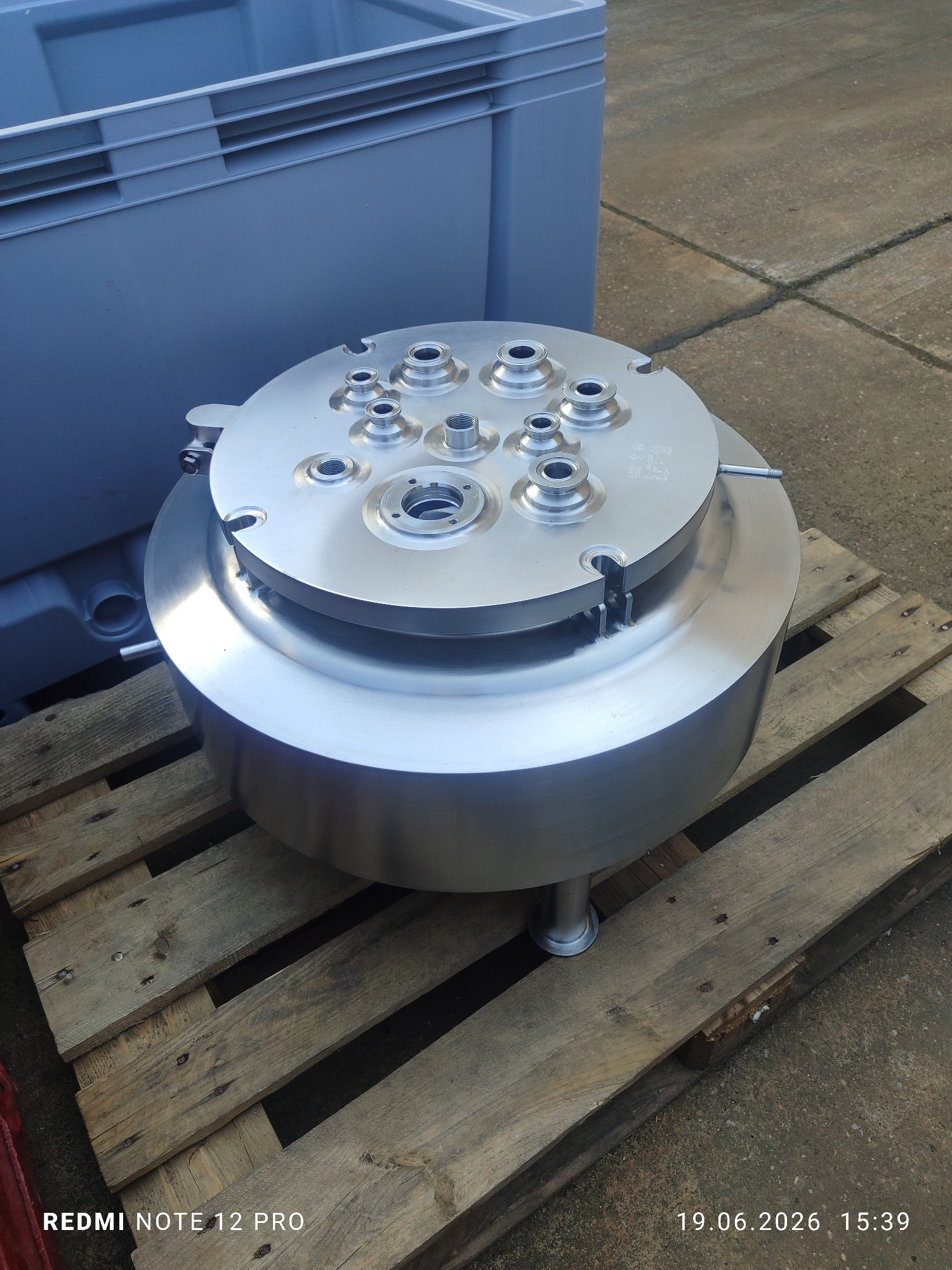 A polished metal cylindrical object with multiple threaded openings on its top surface sits on a wooden pallet. The object is supported by three cylindrical metal legs. In the background, a large, gray plastic container with ribbed sides is visible. The scene is outdoors, on a concrete surface with visible cracks. The timestamp in the lower right corner reads "19.06.2026 15:39". The text "REDMI NOTE 12 PRO" is in the lower left corner. The image was taken in Arnstadt, Germany.