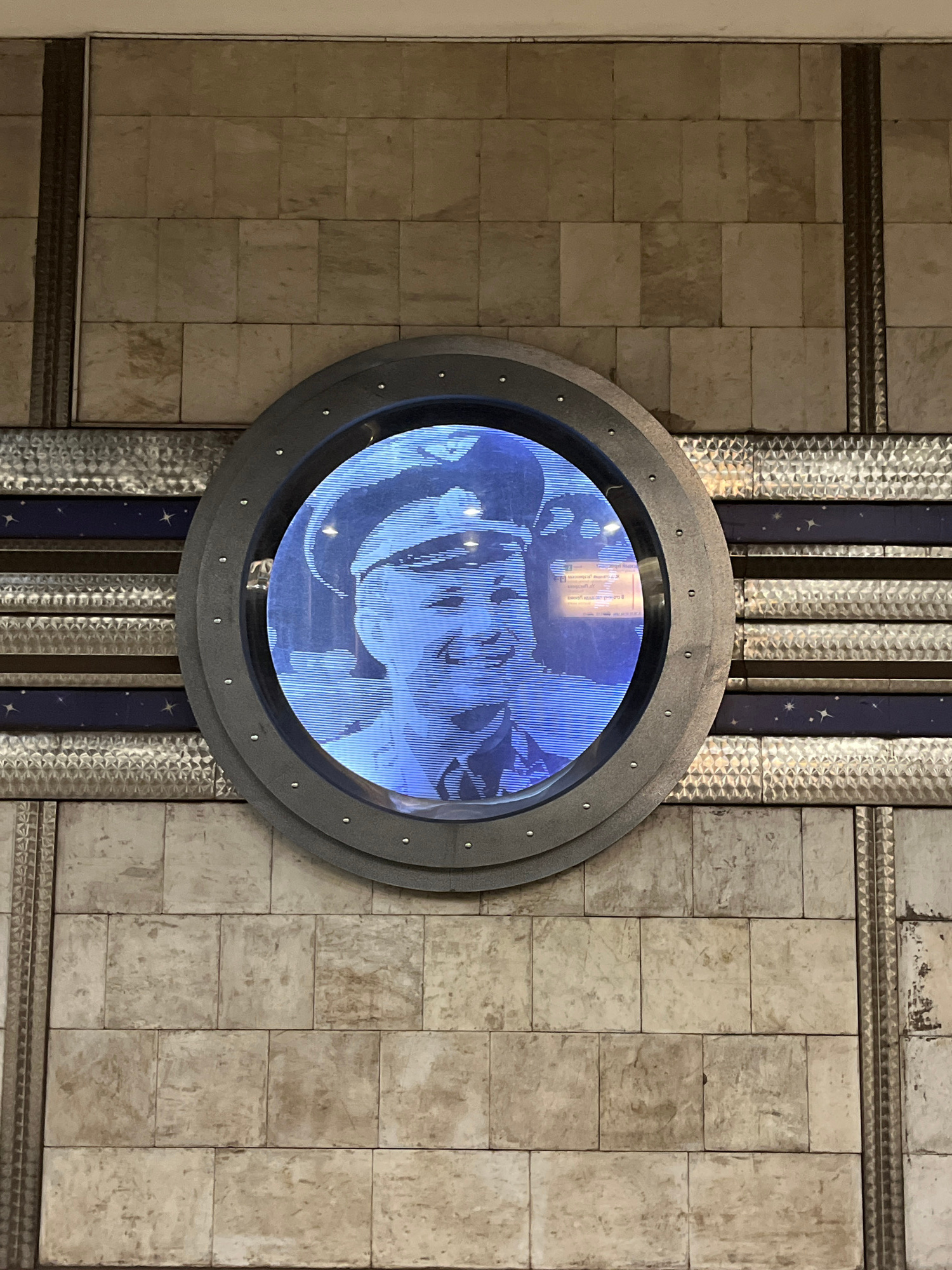 A circular digital display, framed by a dark gray rim with visible rivets, shows a blue-toned, pixelated image of a smiling man wearing a military-style cap and uniform. The display is mounted on a wall covered with beige, square tiles. Below the display, a horizontal band with a metallic, textured surface is interrupted by two dark blue stripes adorned with small, white star-like shapes. The wall is further decorated with vertical metallic trim elements. The background is indistinct, suggesting an indoor or underground setting.