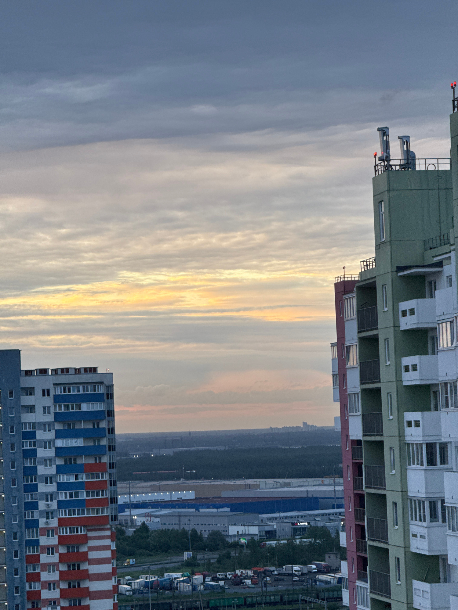 This is a photograph taken from a high vantage point, possibly from another building, looking out over a city landscape at dusk. The sky is filled with layers of clouds, with a strip of pale yellow and orange light indicating the setting sun visible in the middle distance.
In the foreground, two multi-story apartment buildings dominate the scene. The building on the left is tall and slender, with a distinctive facade featuring alternating bands of white, red, and blue on its balconies and walls. It appears to be a residential building. The building on the right is wider and taller, painted in sections of green and white, with protruding balconies. The upper portion of this building features a concrete structure with what appear to be ventilation shafts or equipment, topped with small red warning lights.
Beyond the apartment buildings, the middle ground reveals a sprawling urban and industrial area. There are large, flat-roofed buildings, likely warehouses or industrial facilities, some with visible signage. A dense line of trees separates this industrial zone from the apartment buildings. In the far distance, a hazy cityscape is visible, with the silhouettes of larger buildings and structures against the twilight sky. The presence of trucks and shipping containers near the ground suggests a logistics or industrial hub.
The overall impression is one of a modern urban environment during the transitional hours of evening, with a focus on residential and industrial architecture. The location is identified as Unknown, Russia, based on the provided context.