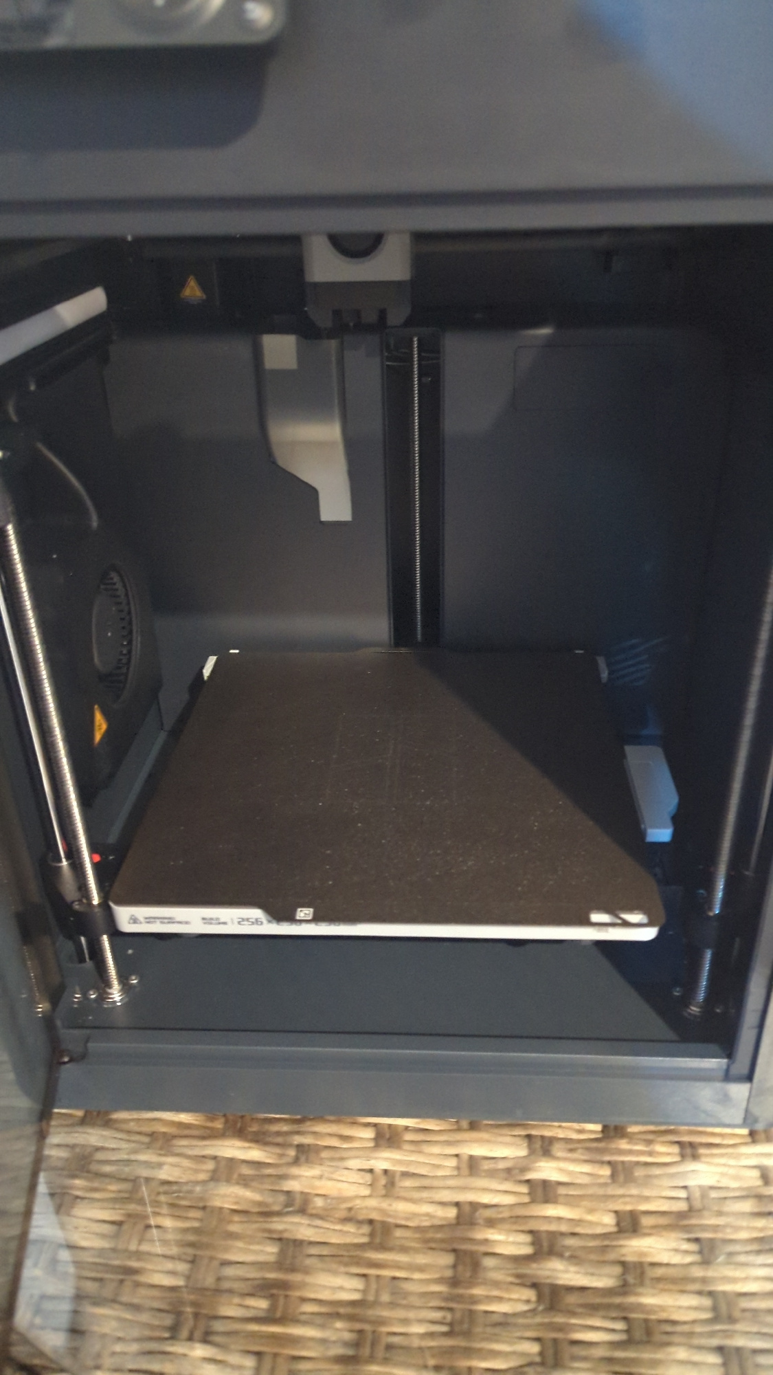 An indoor, eye-level, medium shot captures a 3D printer's build platform and its immediate surroundings. The build platform, a rectangular surface covered with a dark, textured material, is centrally positioned and appears slightly dusty. A white frame with some text and a QR code is visible along the front edge of the platform. The build platform is supported by vertical metal rods and a mechanism that suggests it is part of a larger mechanical system. To the left of the platform, a black fan with a circular grille is mounted on a dark gray surface, and a yellow triangle warning symbol is visible on this surface. The background consists of dark gray and black interior walls of the 3D printer enclosure. The bottom portion of the image shows a textured surface, possibly a woven mat or furniture, rendered in a light brown color with a basket-weave pattern. The lighting is even and artificial, typical of an indoor setting. There are no people or animals present in the image. The scene does not contain any elements that would allow for determining its location in Zadebri, Ukraine.