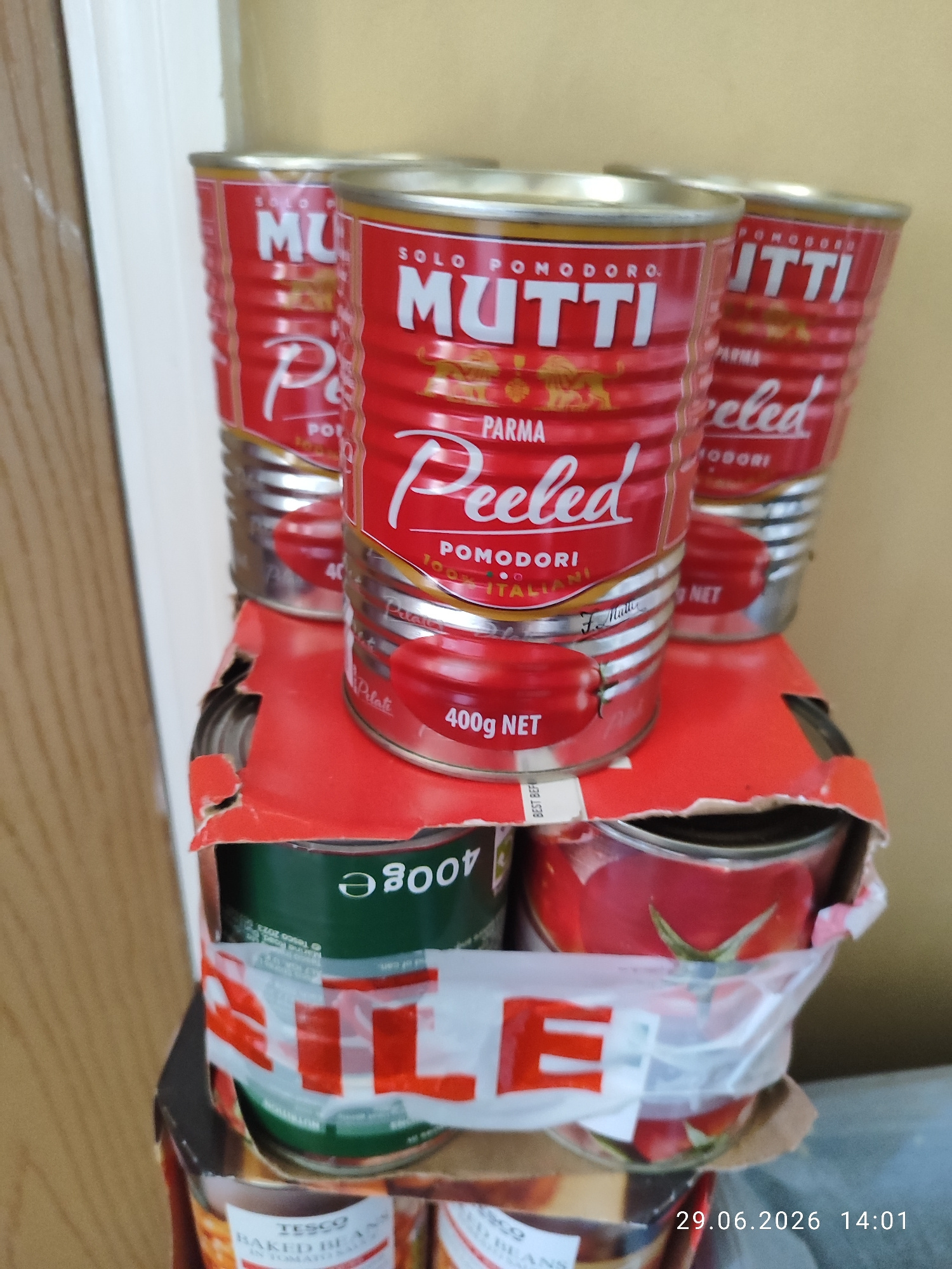 This media file shows a close-up, slightly angled view of several cans of Mutti brand peeled tomatoes stacked in a red cardboard multipack. The cans are primarily red with white and gold lettering. The prominent text on the cans includes "MUTTI," "SOLO POMODORO," "PARMA," and "Peeled POMODORI." One can also clearly shows "400g NET."
Below the top layer of tomato cans, parts of other cans are visible, including a green can with "400g e" in white text, and a can with a red tomato illustration. Further down, a can with "TESCO BAKED BEANS" is partially visible.
The setting appears to be indoors, likely a pantry or storage area, with a wall of light-colored wood or laminate visible on the left and a pale yellow wall in the background. The lighting suggests it is daytime. A timestamp in the bottom right corner reads "29.06.2026 14:01," indicating the photo was taken on June 29, 2026, at 2:01 PM. The overall impression is one of food items being stored or packaged. There are no people or animals visible in the image.